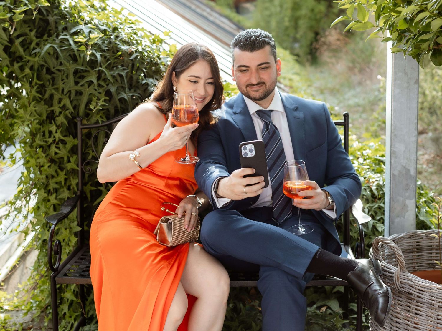 Couple in formal attire enjoying cocktails together on a garden bench at an outdoor wedding reception.