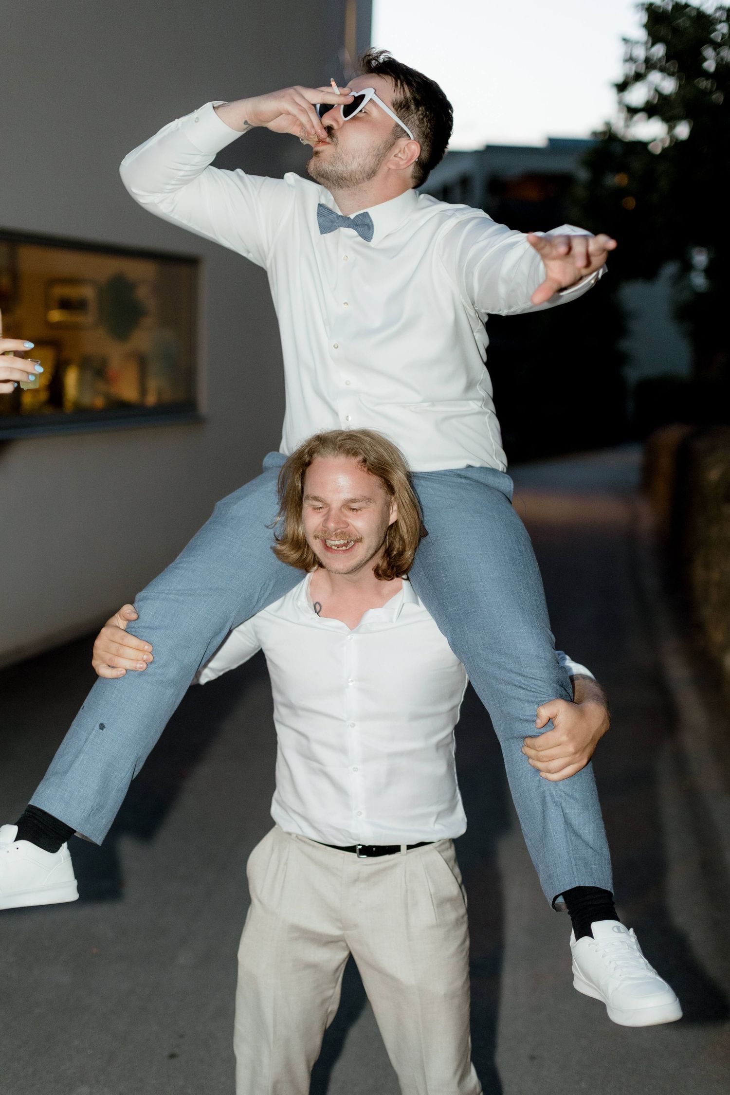 Man in heart sunglasses riding on friends shoulders while drinking at an outdoor wedding.