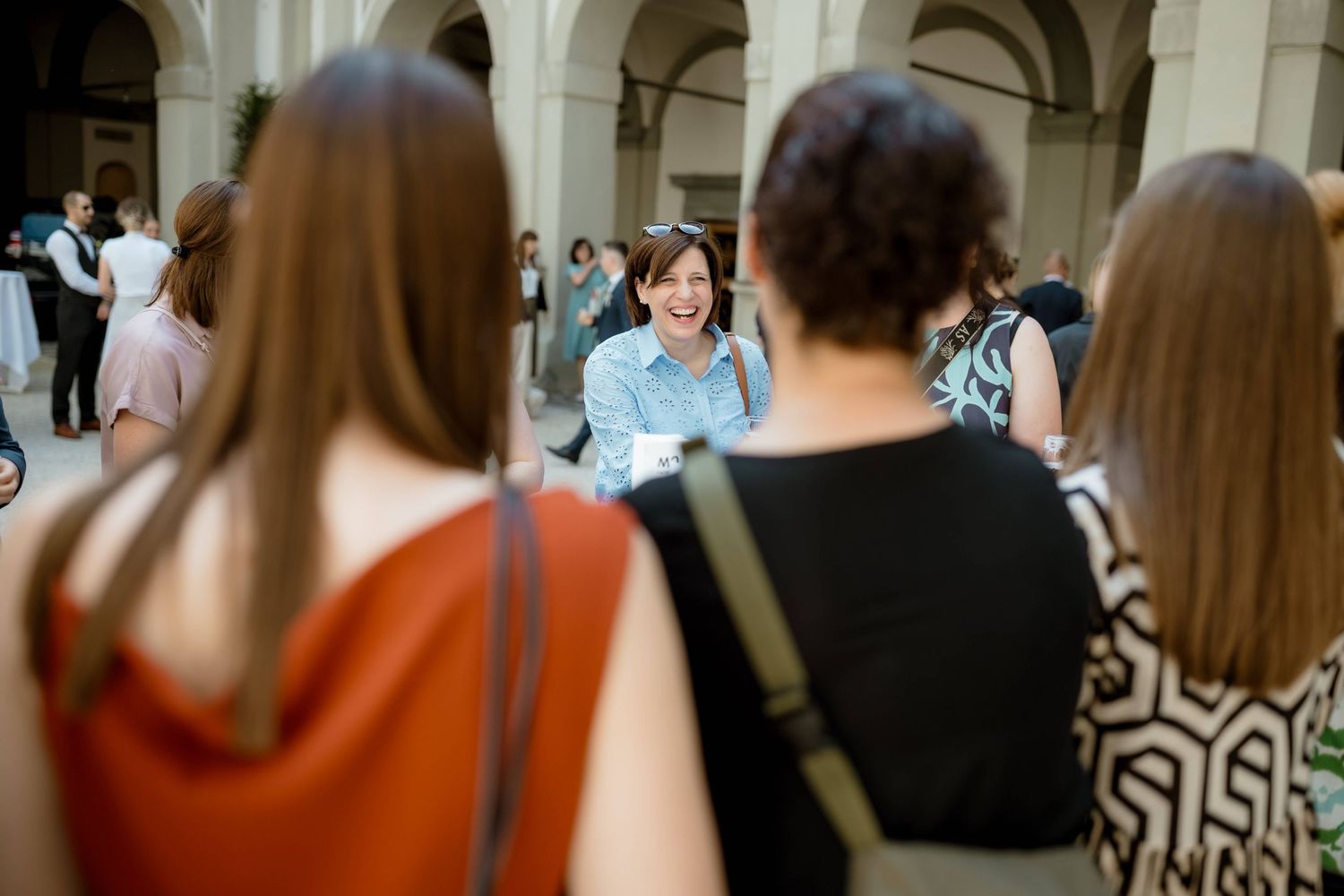 Smiling woman in denim jacket chatting with guests at an outdoor wedding gathering in a courtyard.