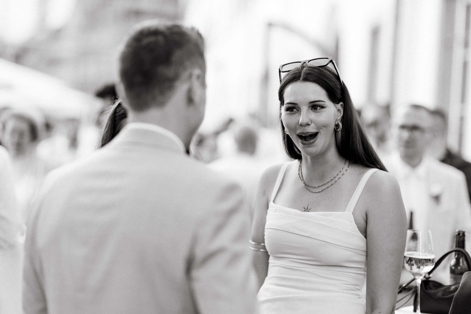 Elegant woman in white dress with sunglasses smiles at man in black and white photo.