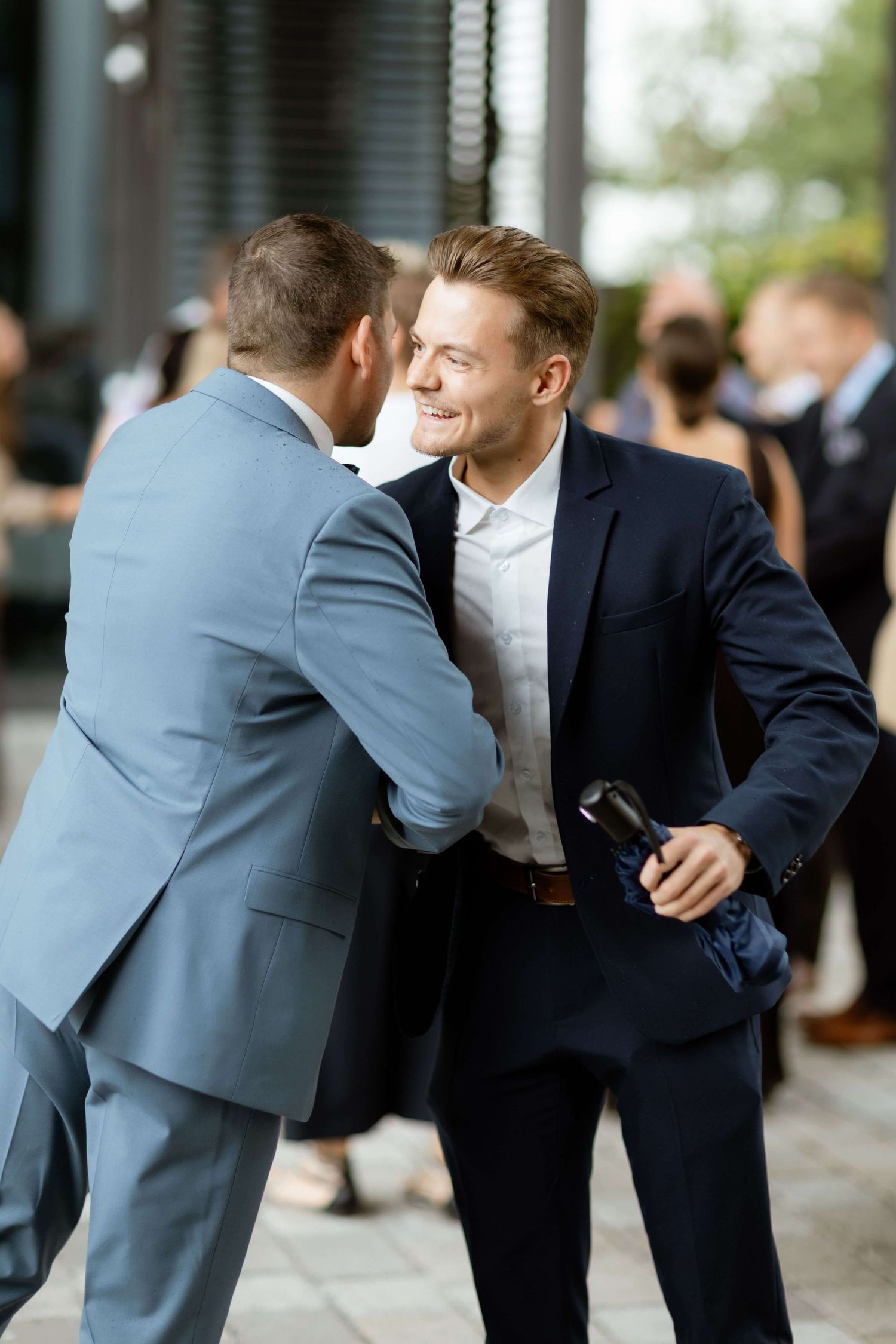 Two men in suits greet each other warmly at an outdoor wedding reception event.