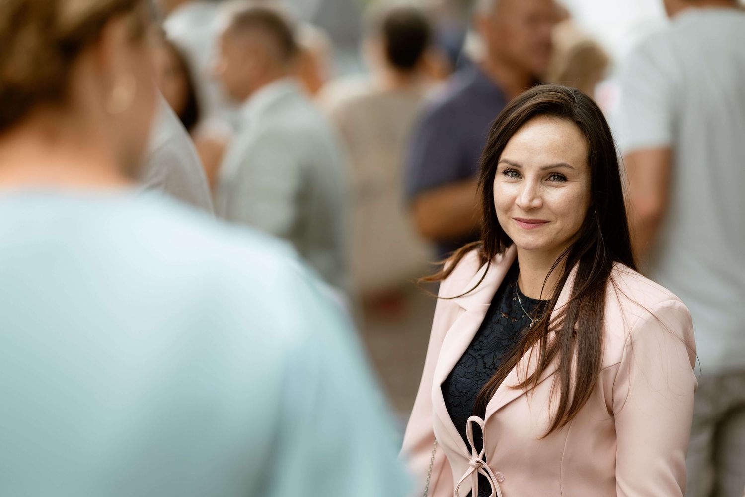 A woman in a pink blazer smiles warmly at someone in a crowded indoor event setting.