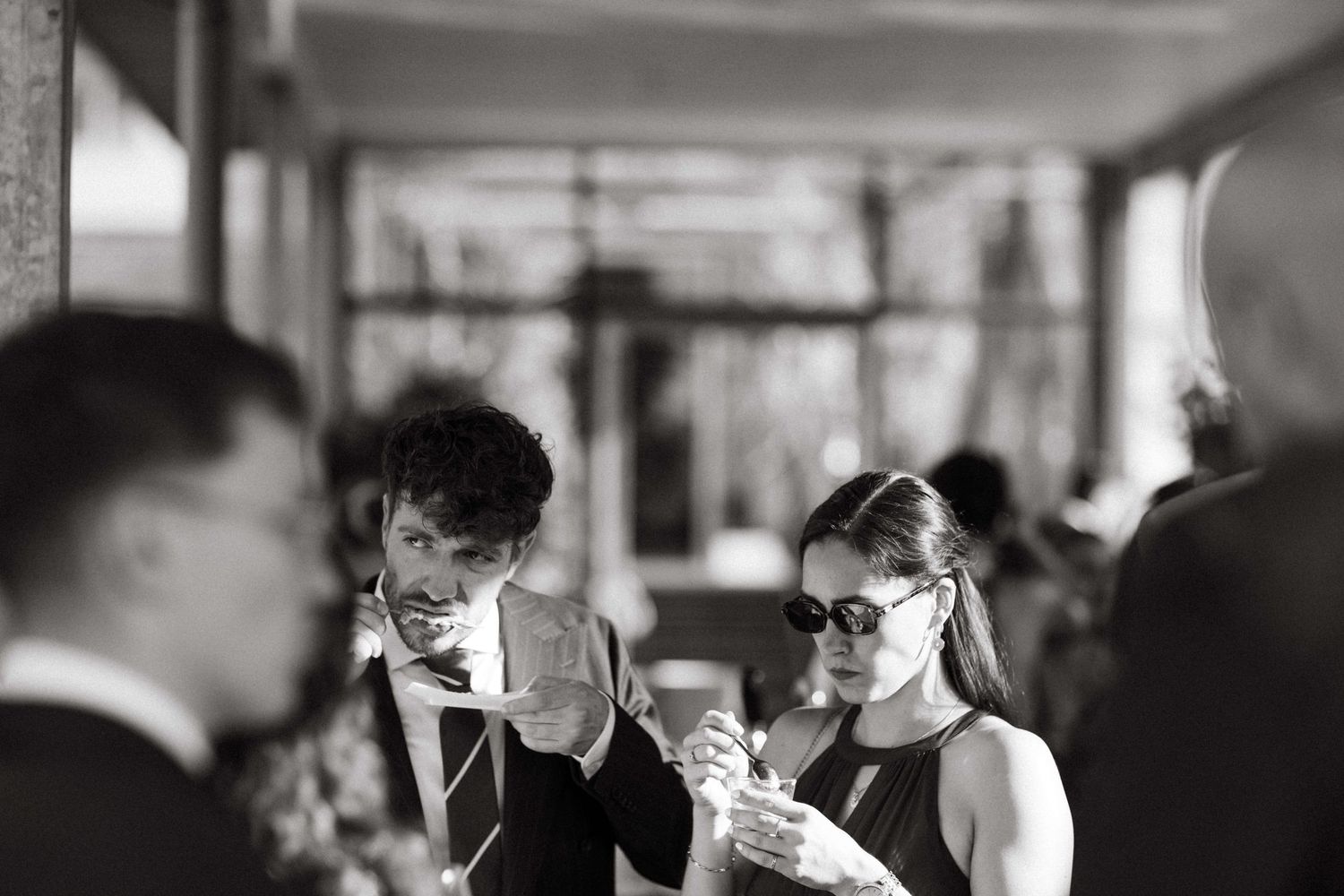 Black and white photo of wedding guests in formal wear looking at phones during an outdoor reception.