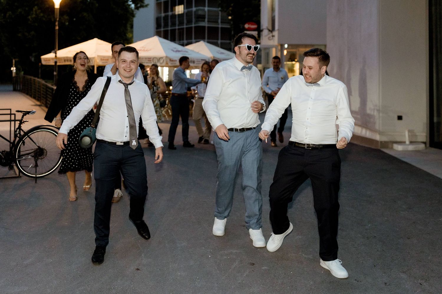 Three men in dress shirts walk and dance playfully on an outdoor plaza during an evening wedding celebration.