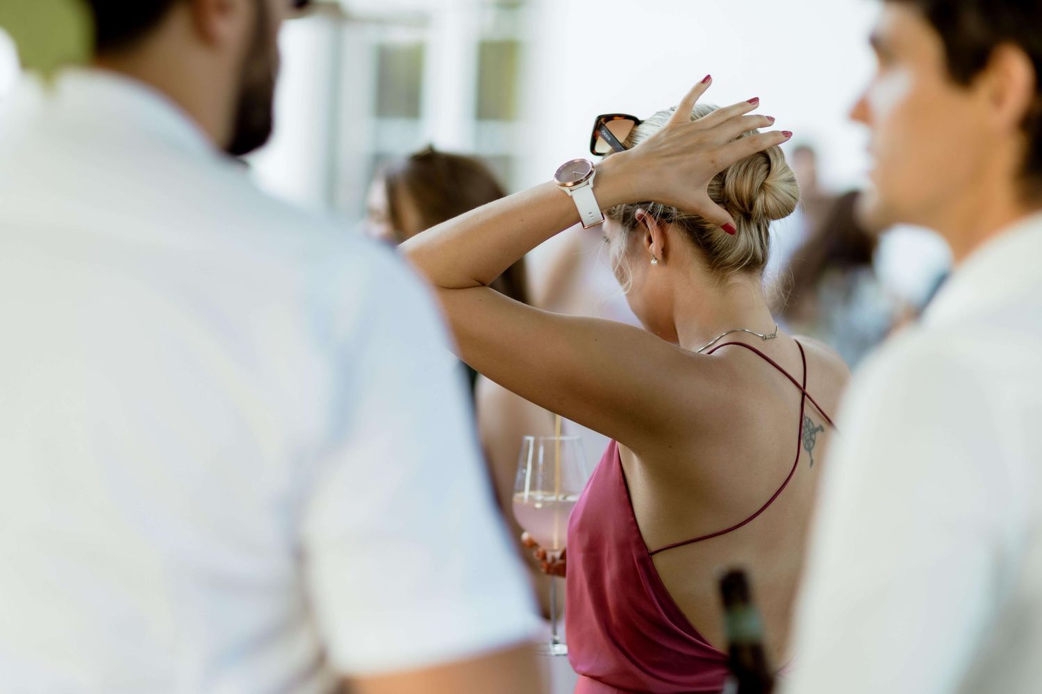Woman in pink dress adjusts hair while holding wine glass at elegant social gathering.