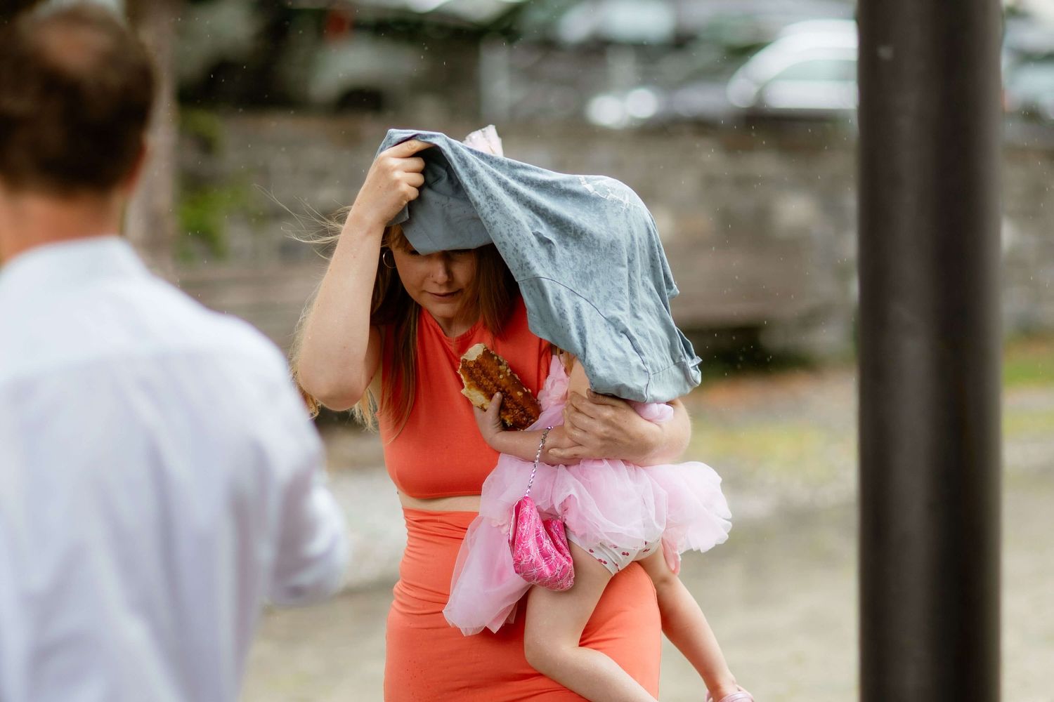 Woman in orange dress shields baby girl in pink tutu from rain using a jacket outdoors.