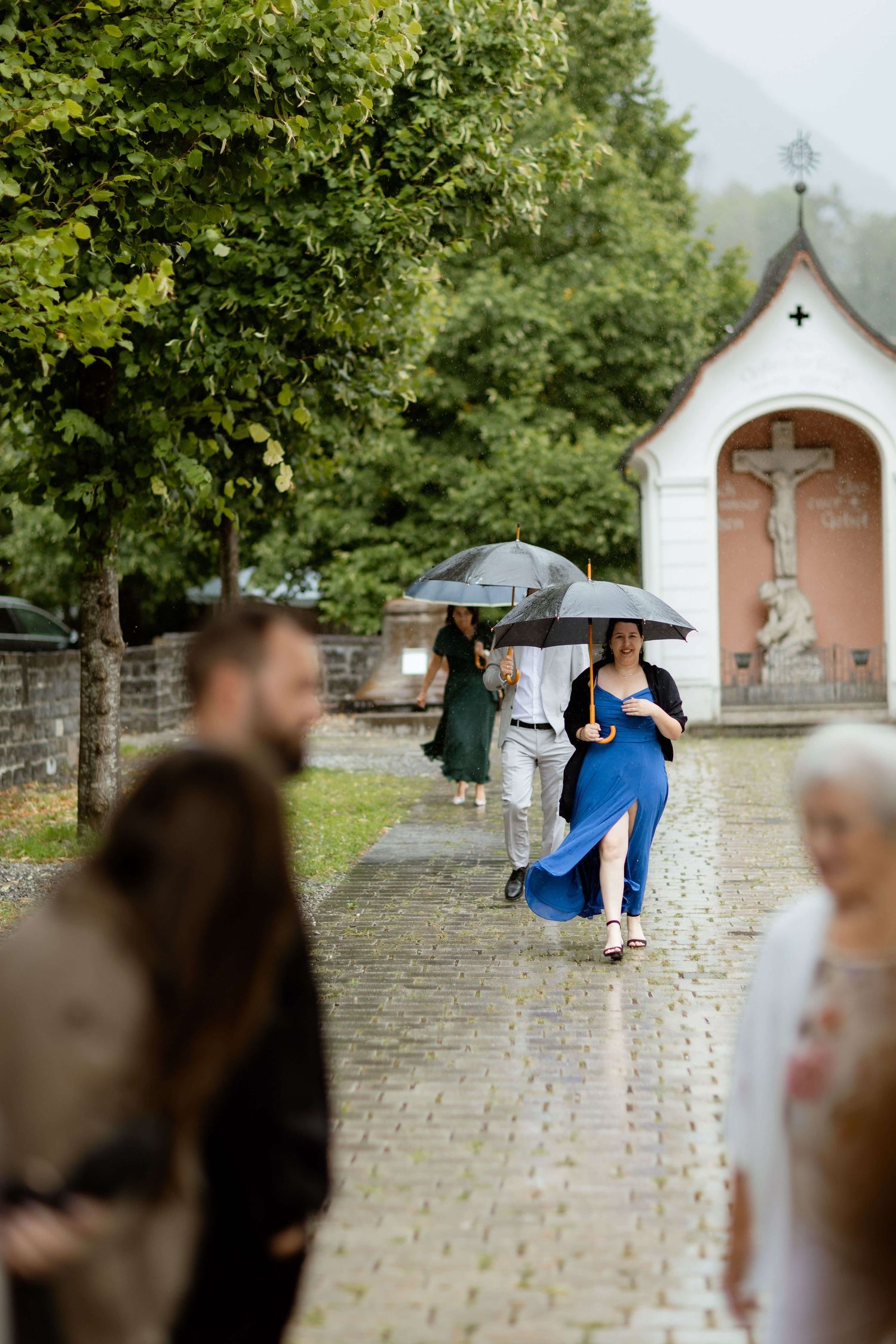 Wedding guests walk with umbrellas along cobblestone path near chapel shrine in the rain.