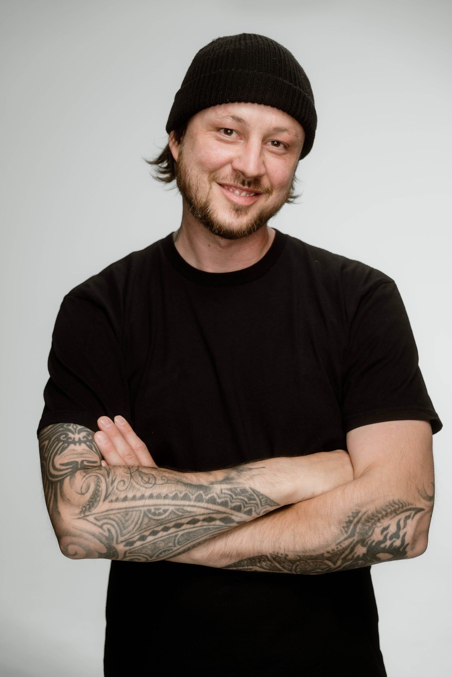Smiling man with tattoos wearing black t-shirt and beanie with arms crossed.
