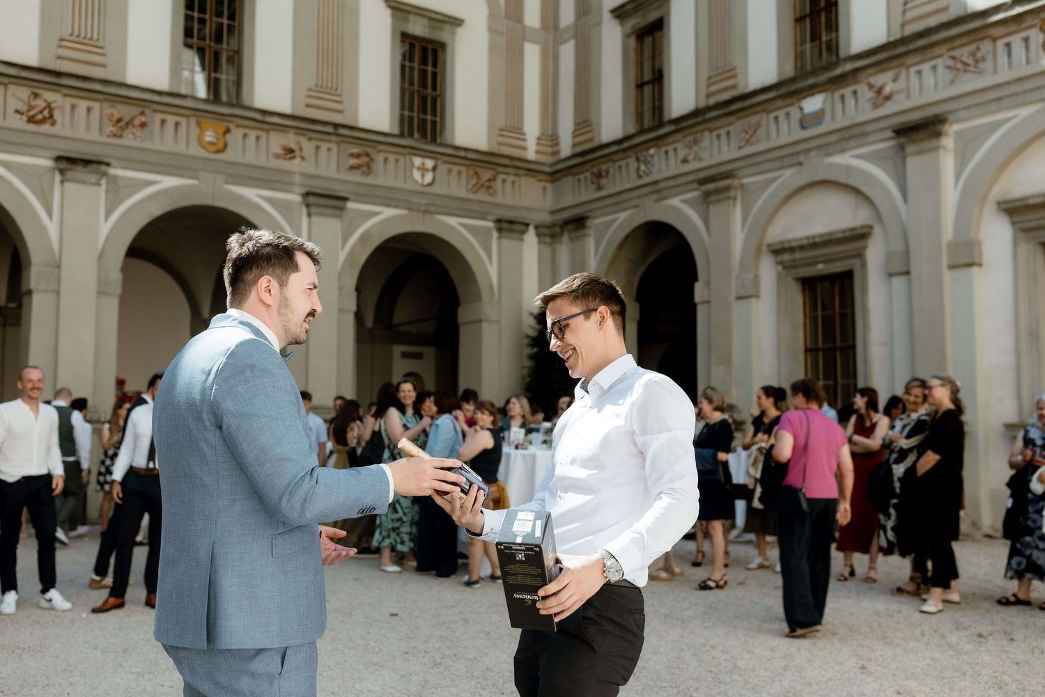Two men in formal attire shaking hands at an outdoor wedding reception in a courtyard.