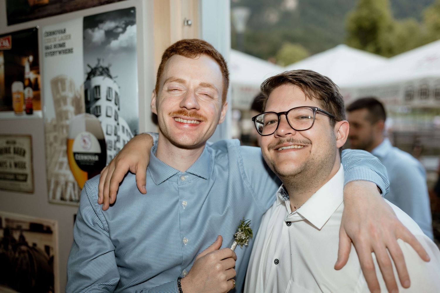Two smiling young men with mustaches pose with arms around each other at an outdoor wedding reception.
