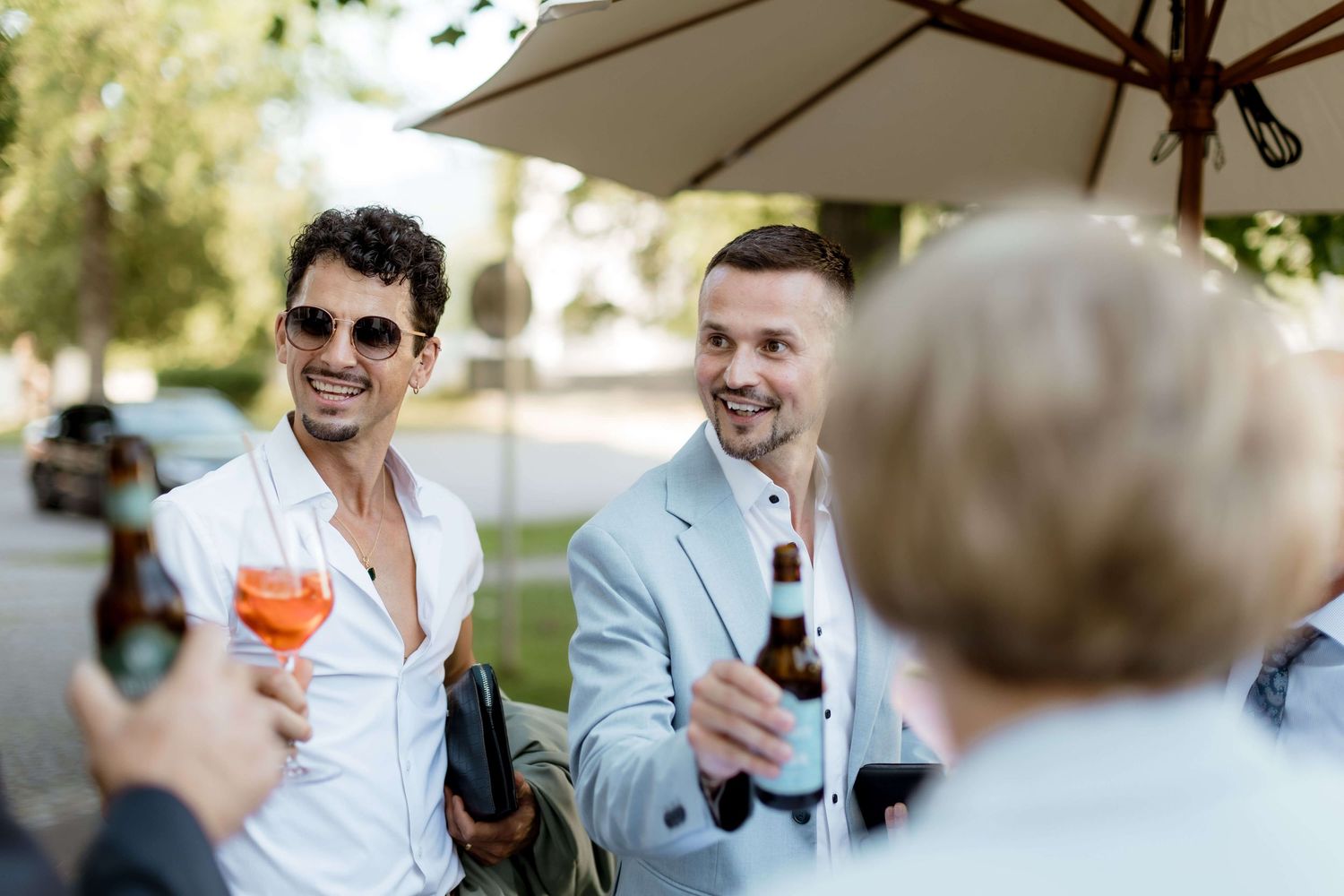 Men in suits socializing outdoors at a wedding reception, holding drinks and smiling under a patio umbrella.