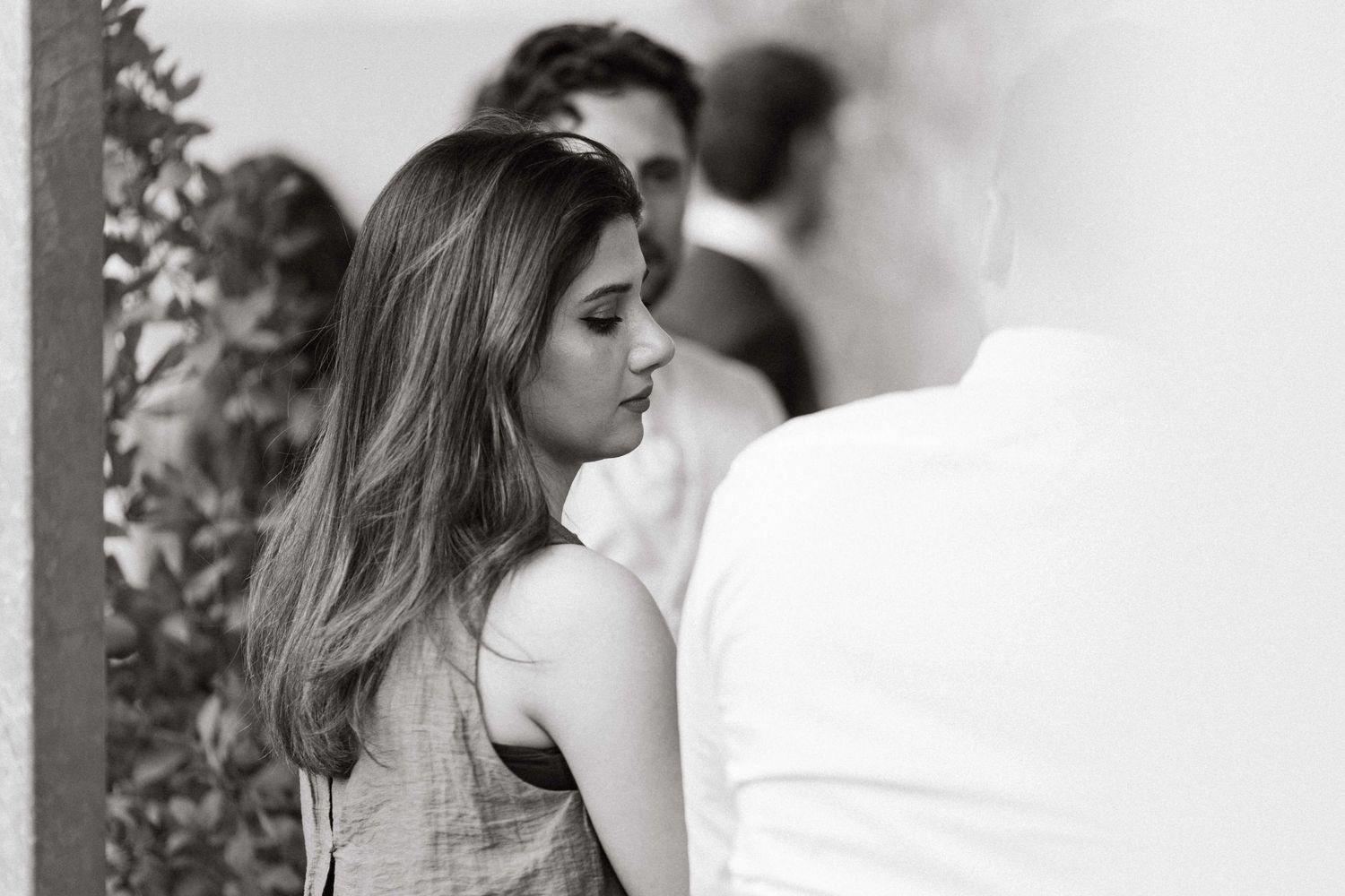 A young woman with long brown hair looks down thoughtfully in a black and white candid photo at a social event.