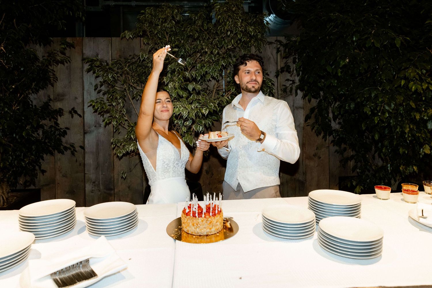 Bride and groom cutting wedding cake outdoors at night with stacked white plates on table.