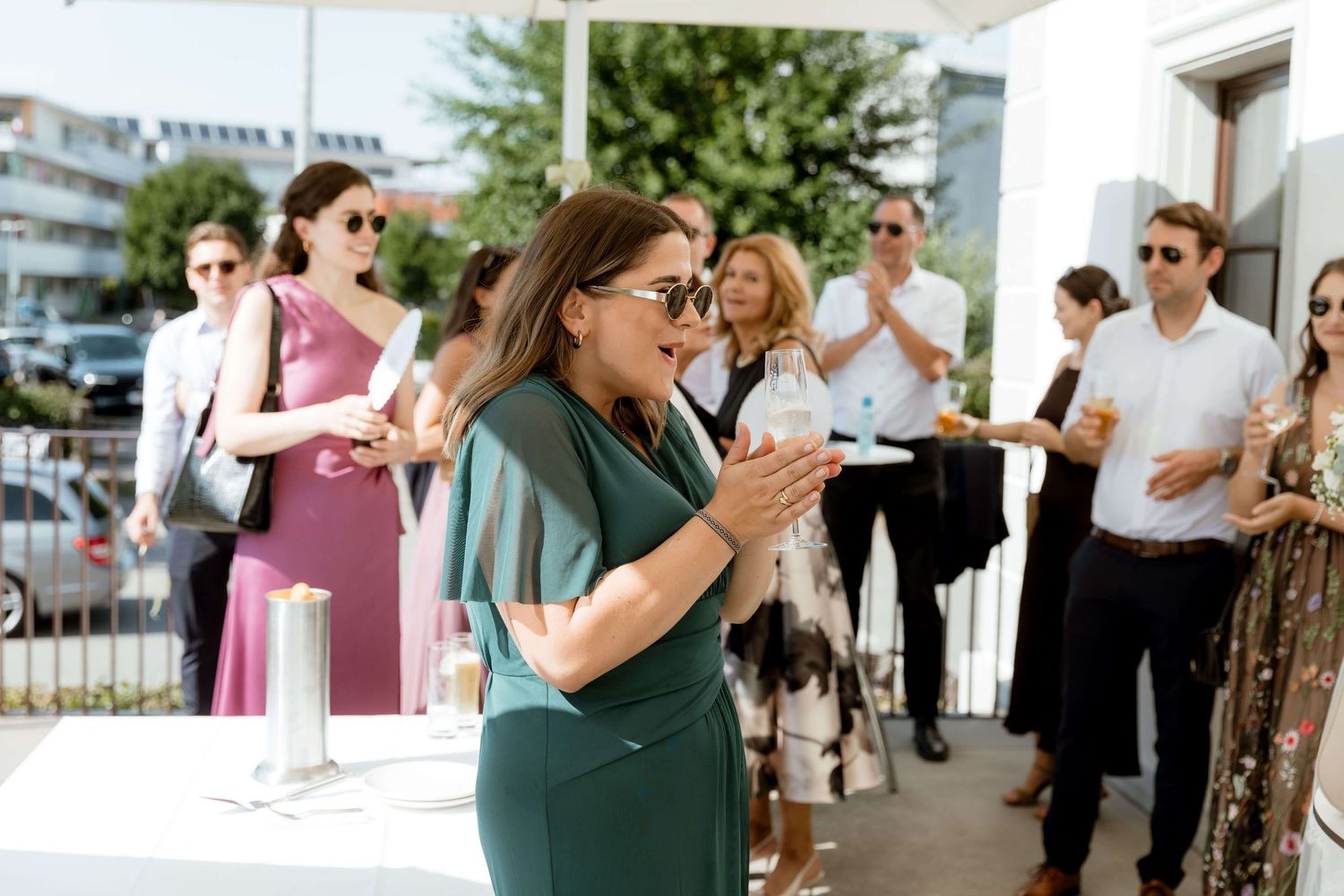 Woman in green dress clapping joyfully while guests celebrate outside at a sunny wedding reception.