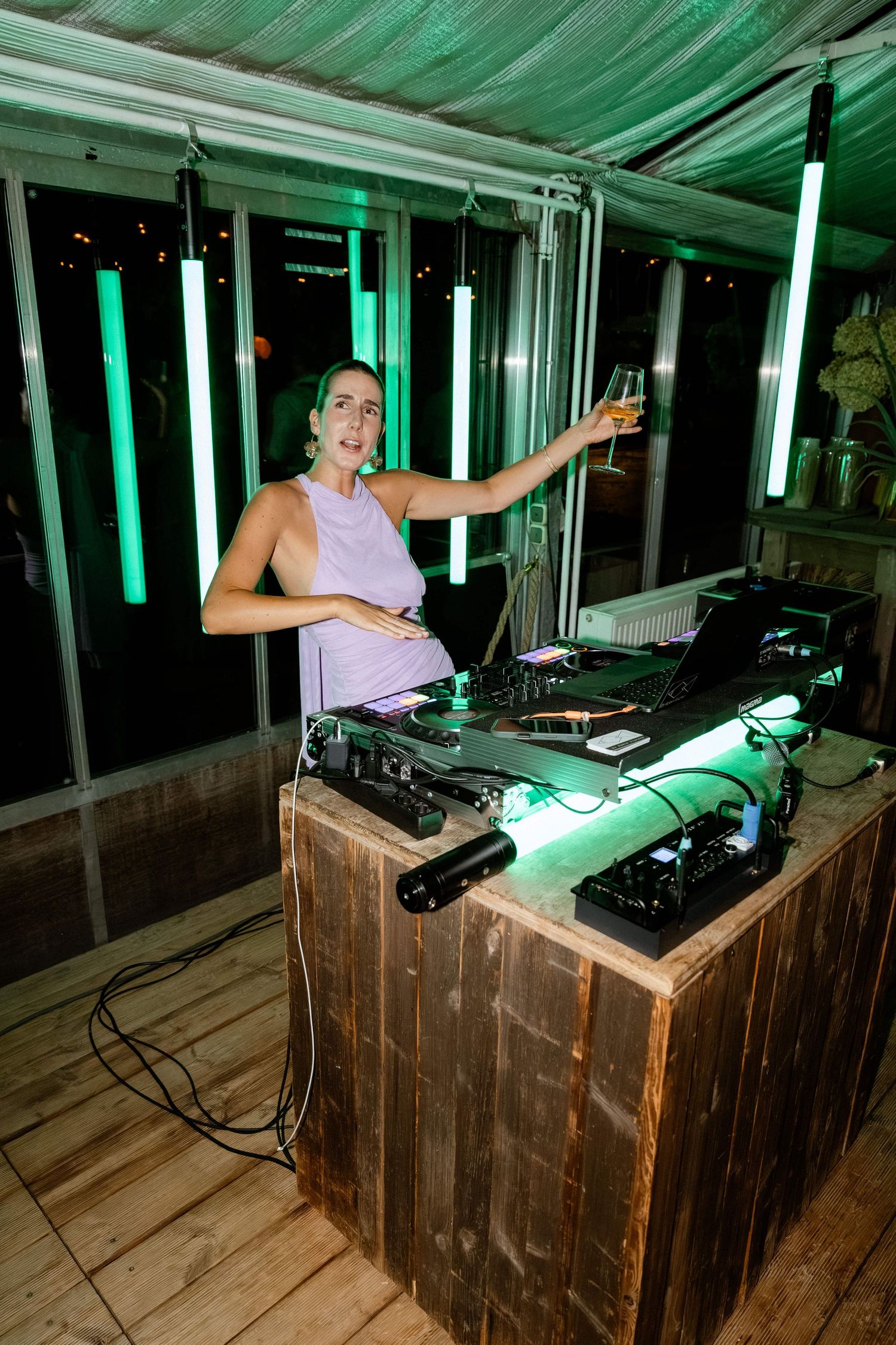 Female DJ in lavender dress raises champagne glass while mixing music at wedding reception.