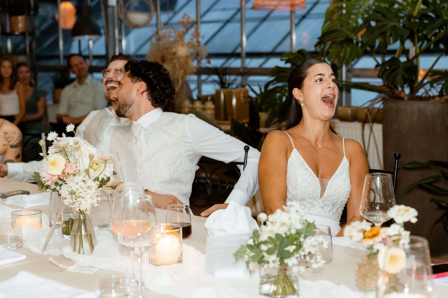 Bride and groom laughing joyfully at wedding reception table with candles and white florals.