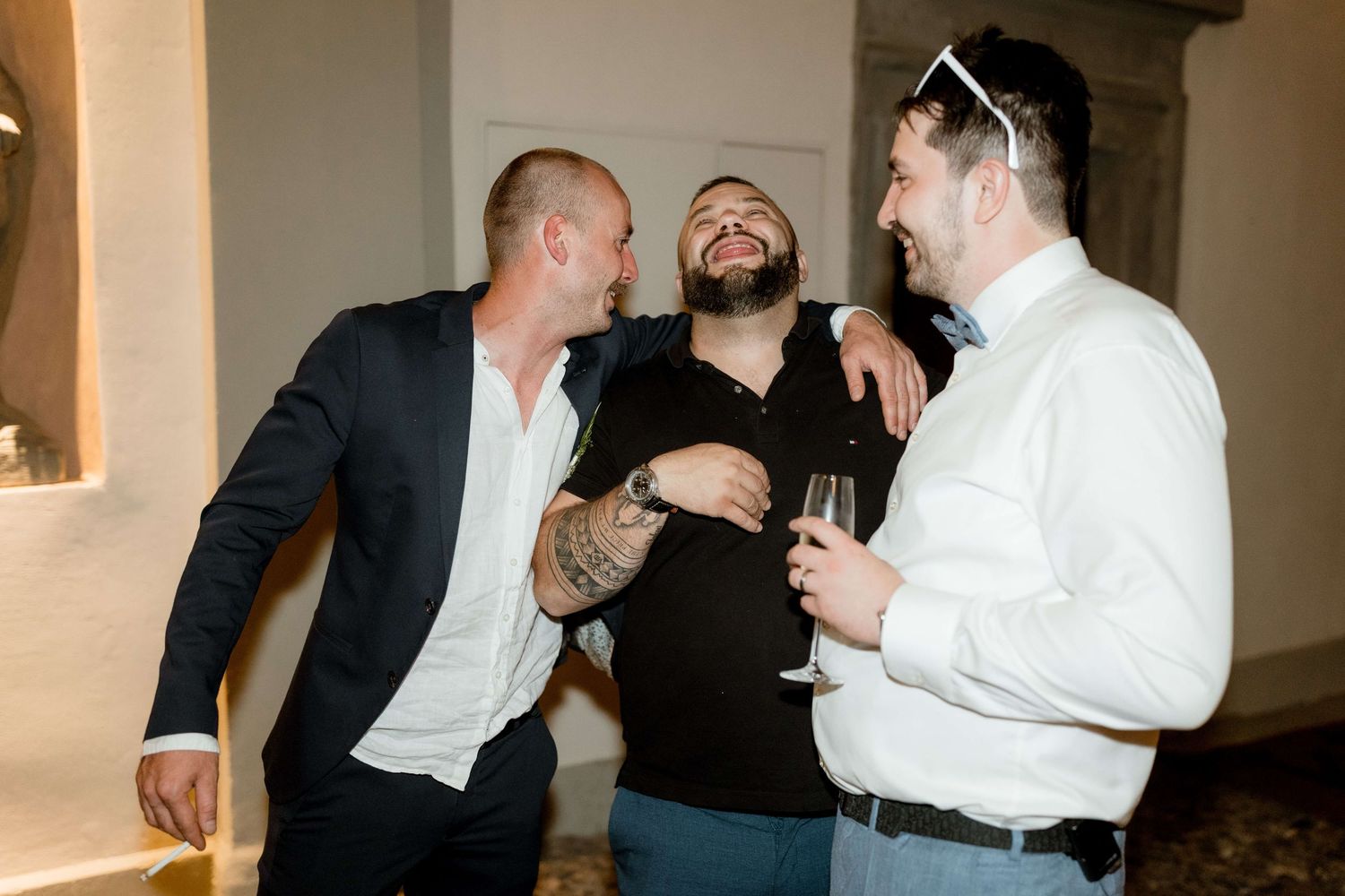 Three men laugh and socialize together while holding champagne glasses at an elegant wedding venue.
