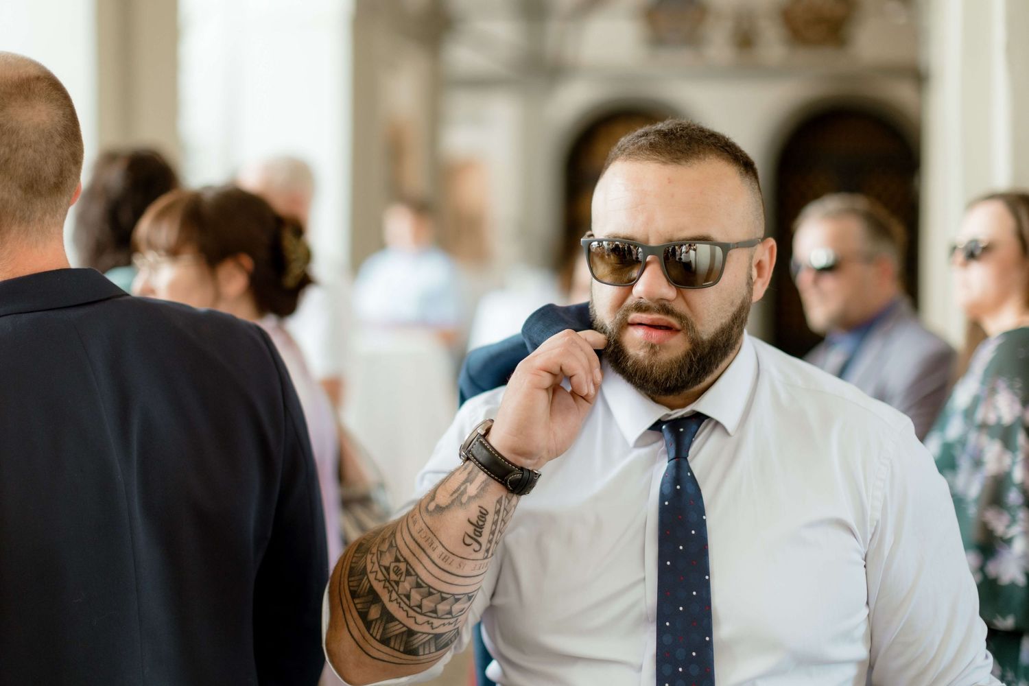 Bearded man with tattoos and sunglasses in white shirt and tie at an outdoor wedding event.