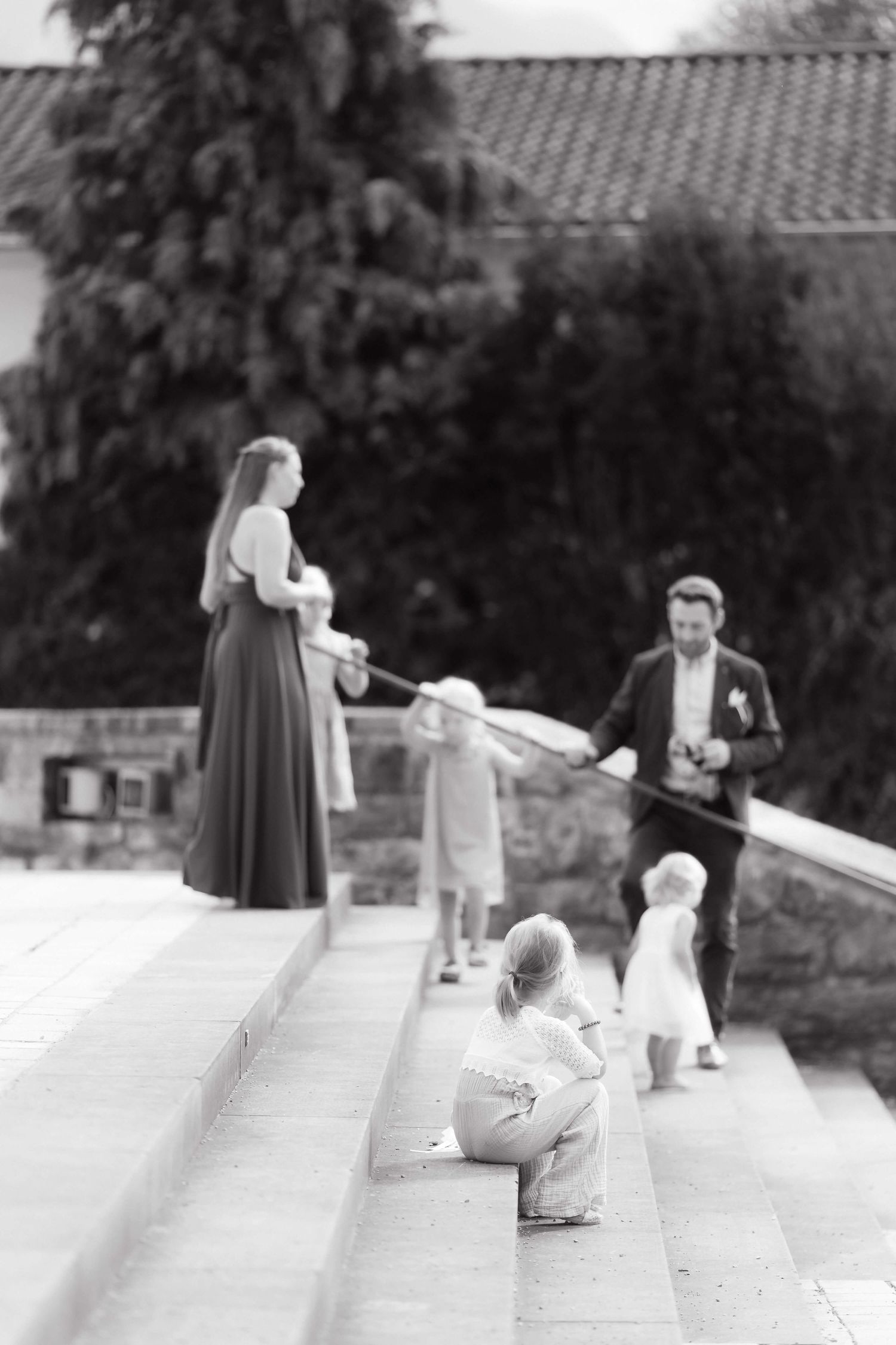Black and white photo of a family on outdoor steps, with children playing and adults in formal attire at a wedding.
