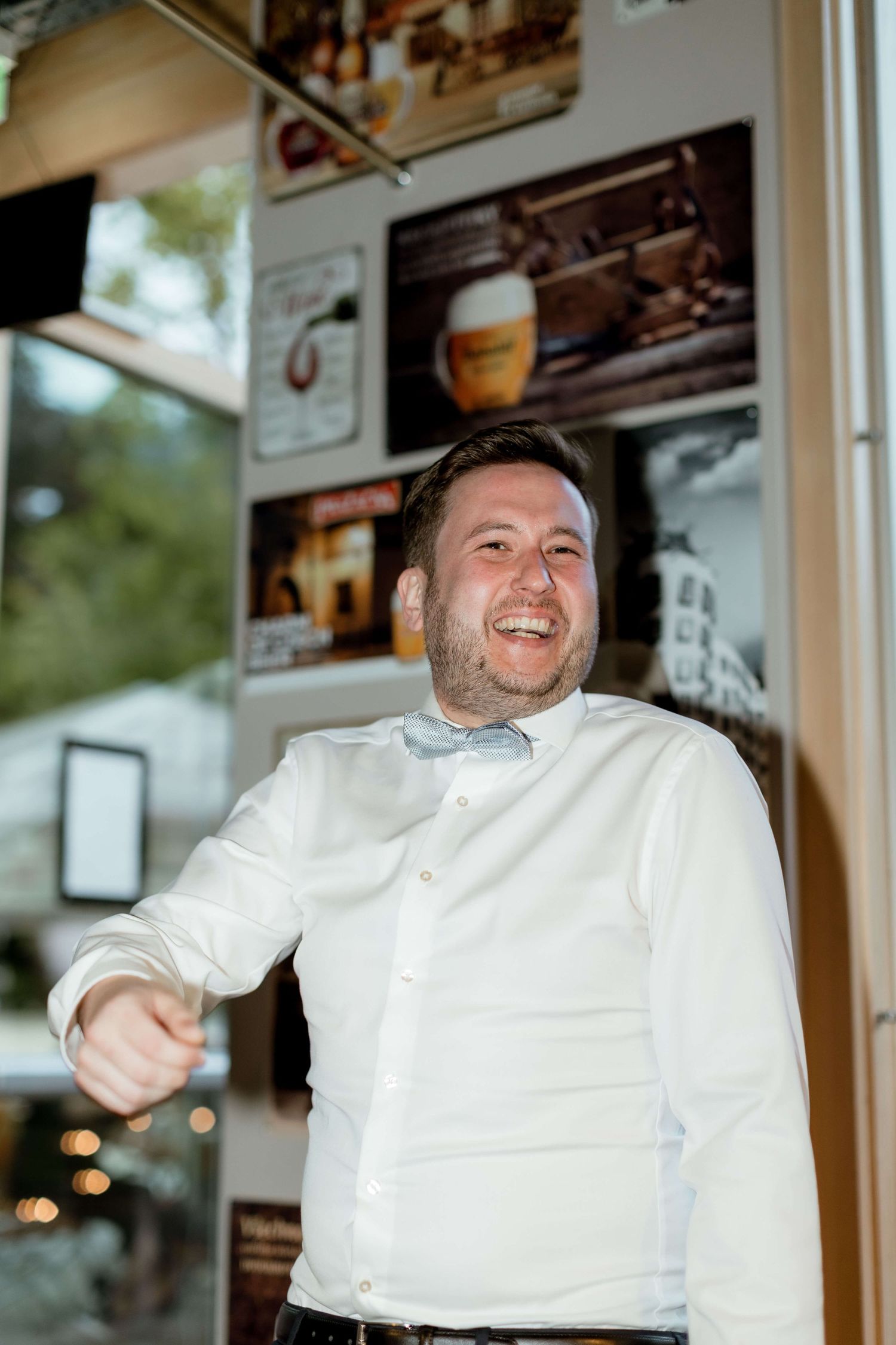 Cheerful man in a white shirt and bow tie laughs while pointing at the camera outside a bar.