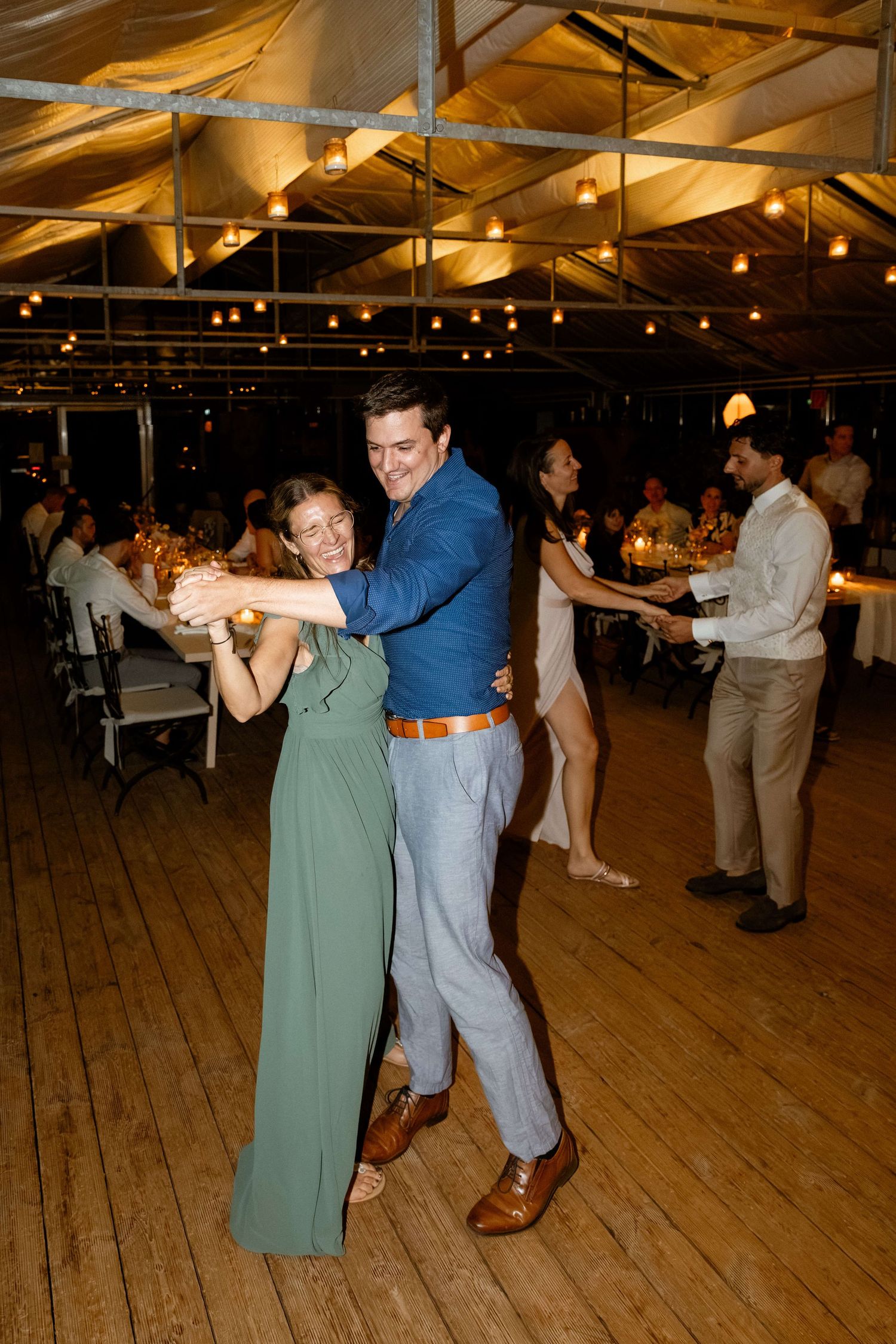 Couple dancing together at wedding reception under string lights on a wooden dance floor.