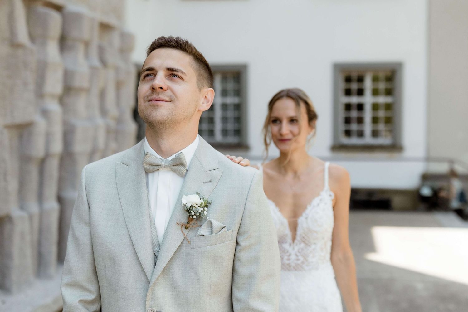 Groom in beige suit looks up while bride in lace wedding dress smiles behind him.