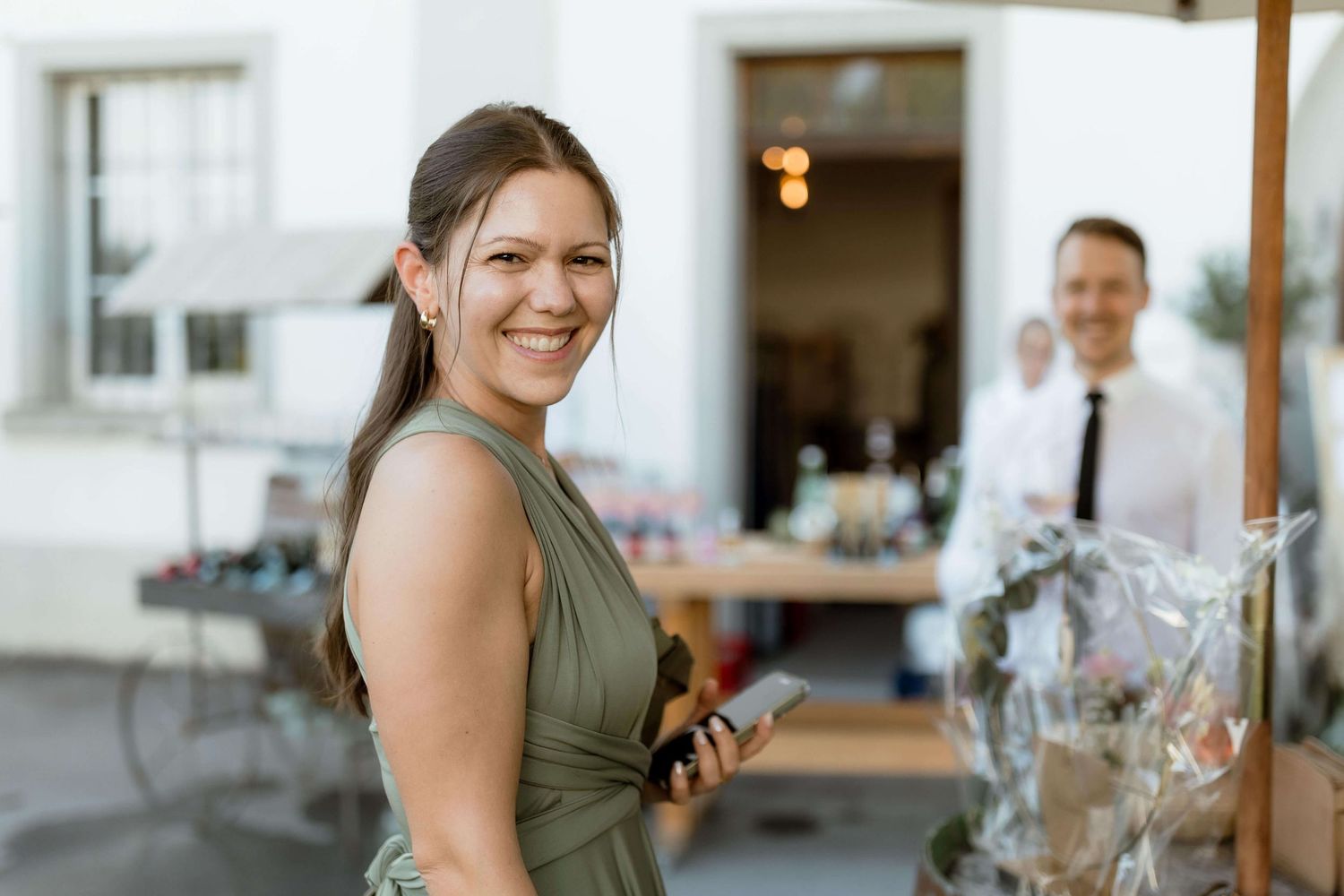 Smiling woman in olive green dress holding phone at outdoor wedding cocktail hour.