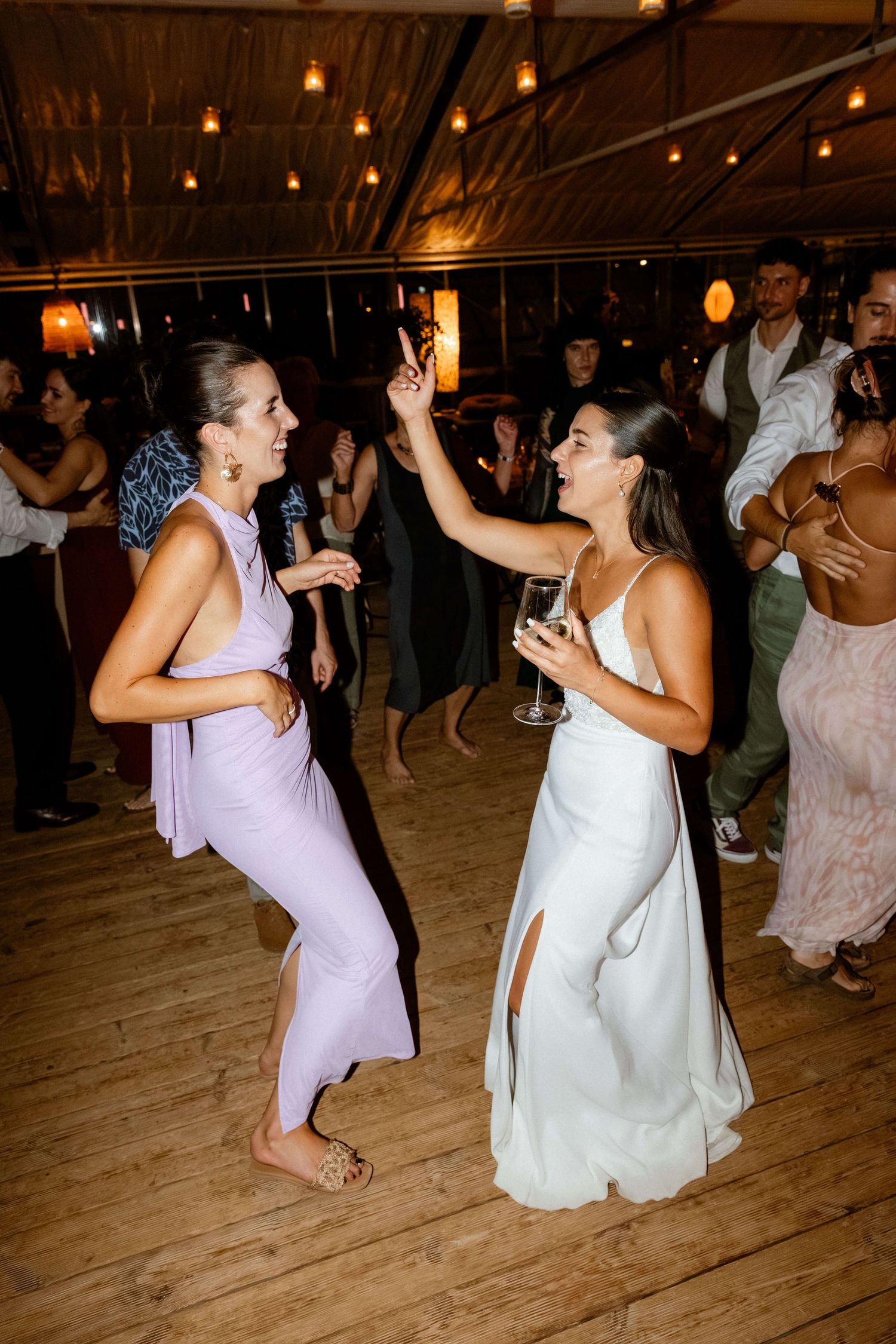 Bride in white dress dancing with friend in lavender gown at lively wedding reception.