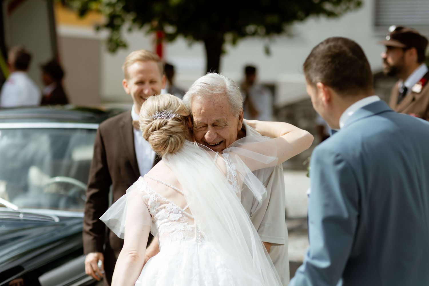 Bride in white gown sharing emotional hug with elderly guest outside church.