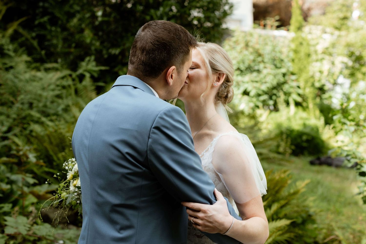 Bride and groom share a romantic kiss surrounded by lush green garden foliage.