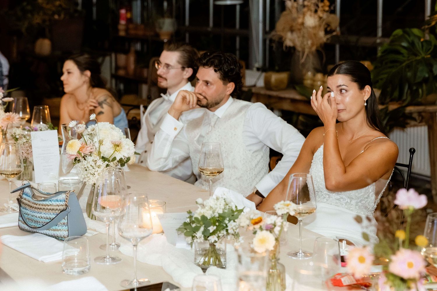 Bride and groom seated at wedding reception table with candles, flowers, and glassware listening to speeches.