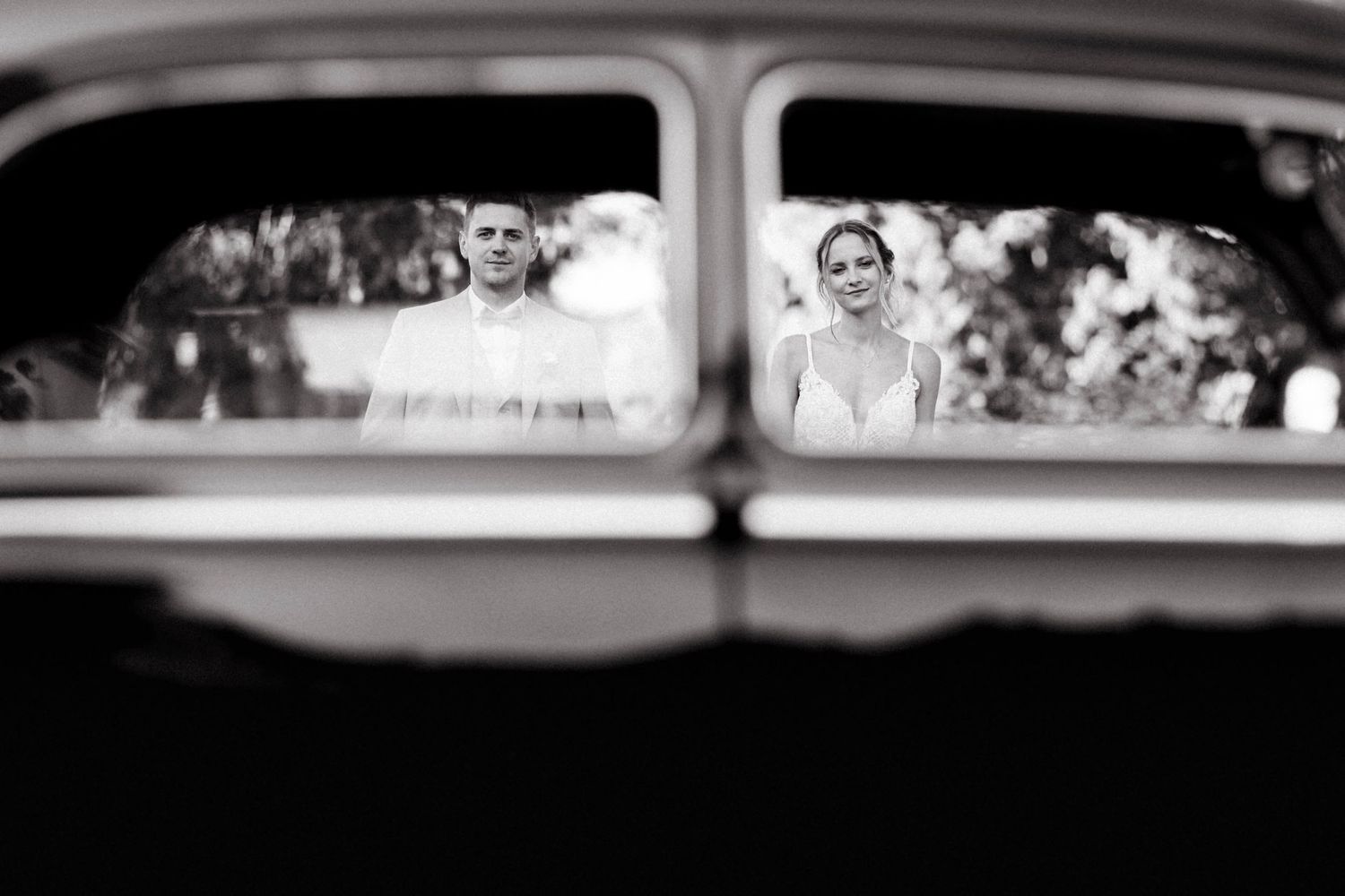 Bride and groom reflected in vintage car window in artistic black and white wedding photo.