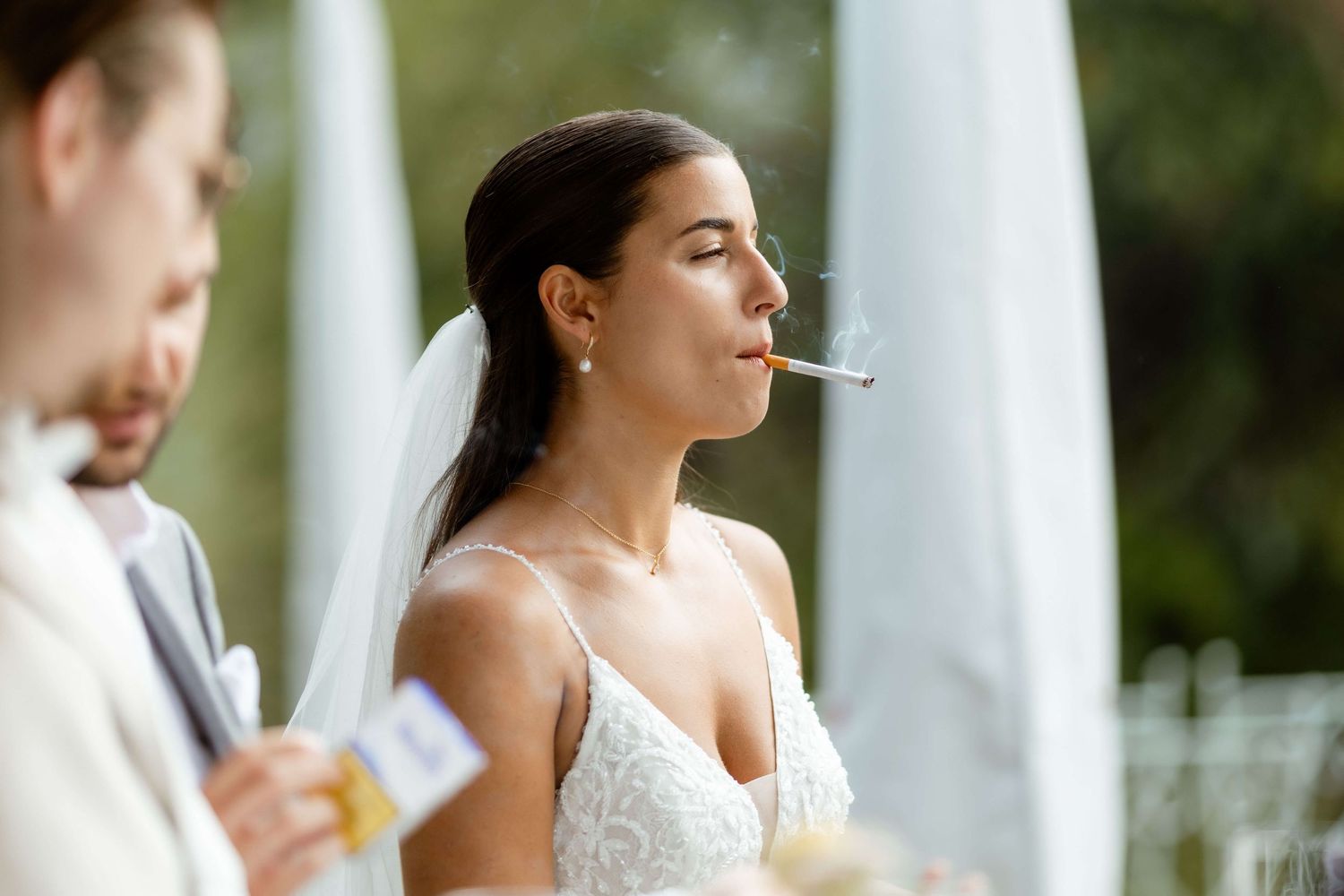 Bride in white lace gown and veil smoking a cigarette during outdoor wedding ceremony.