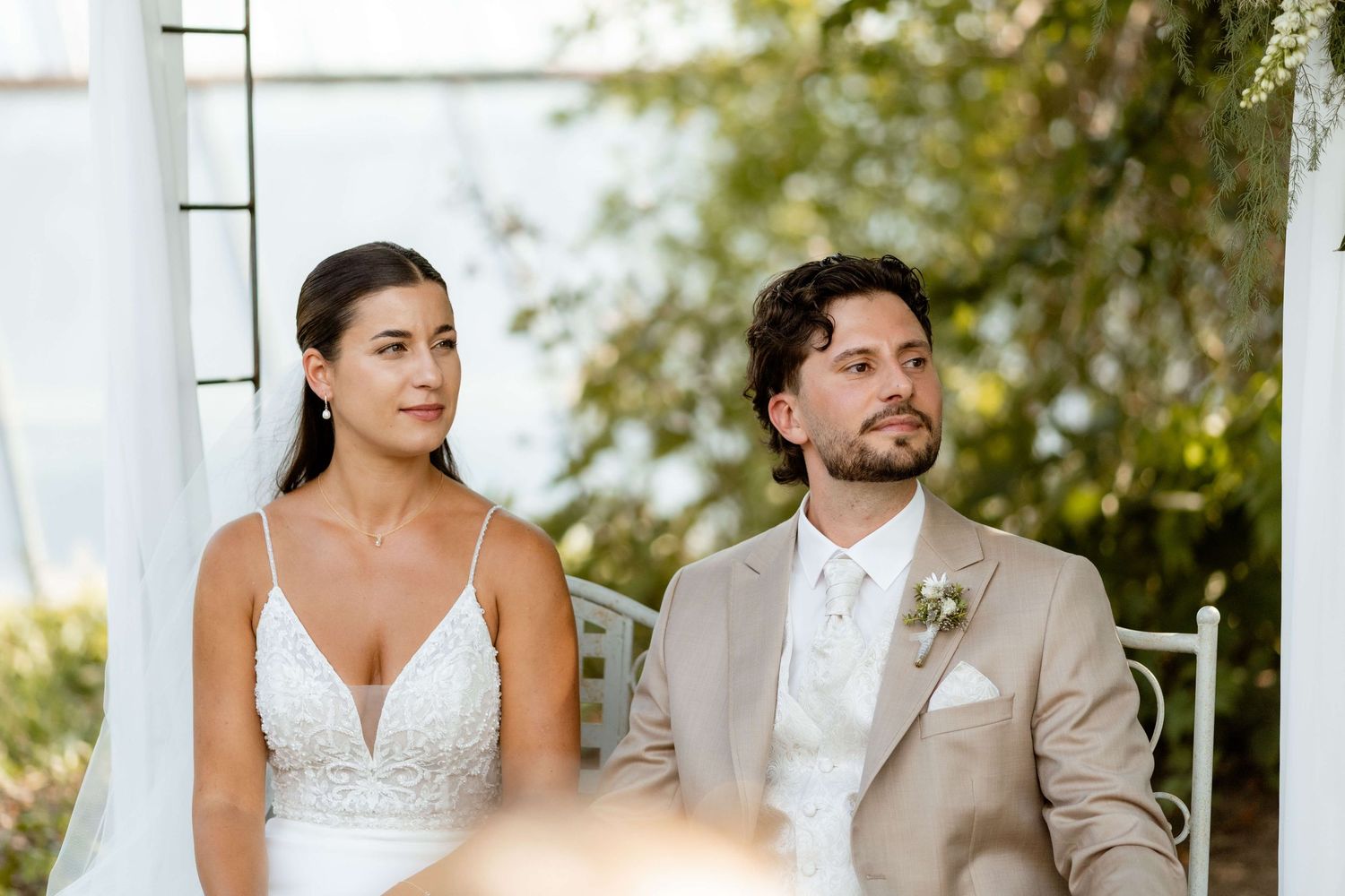 Bride in white gown and groom in tan suit seated together during outdoor wedding ceremony.