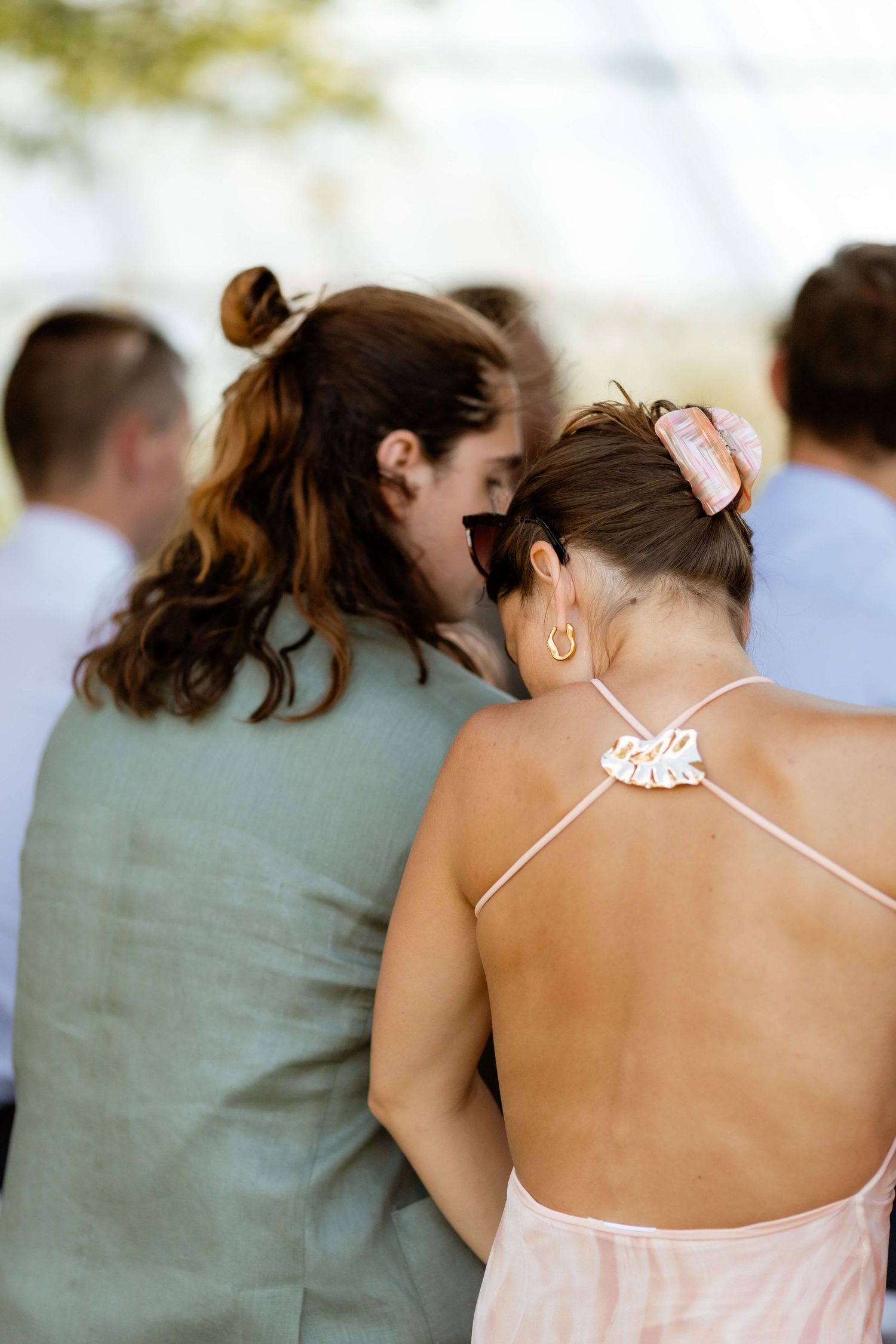 Two female wedding guests lean heads together sharing a moment during outdoor ceremony.