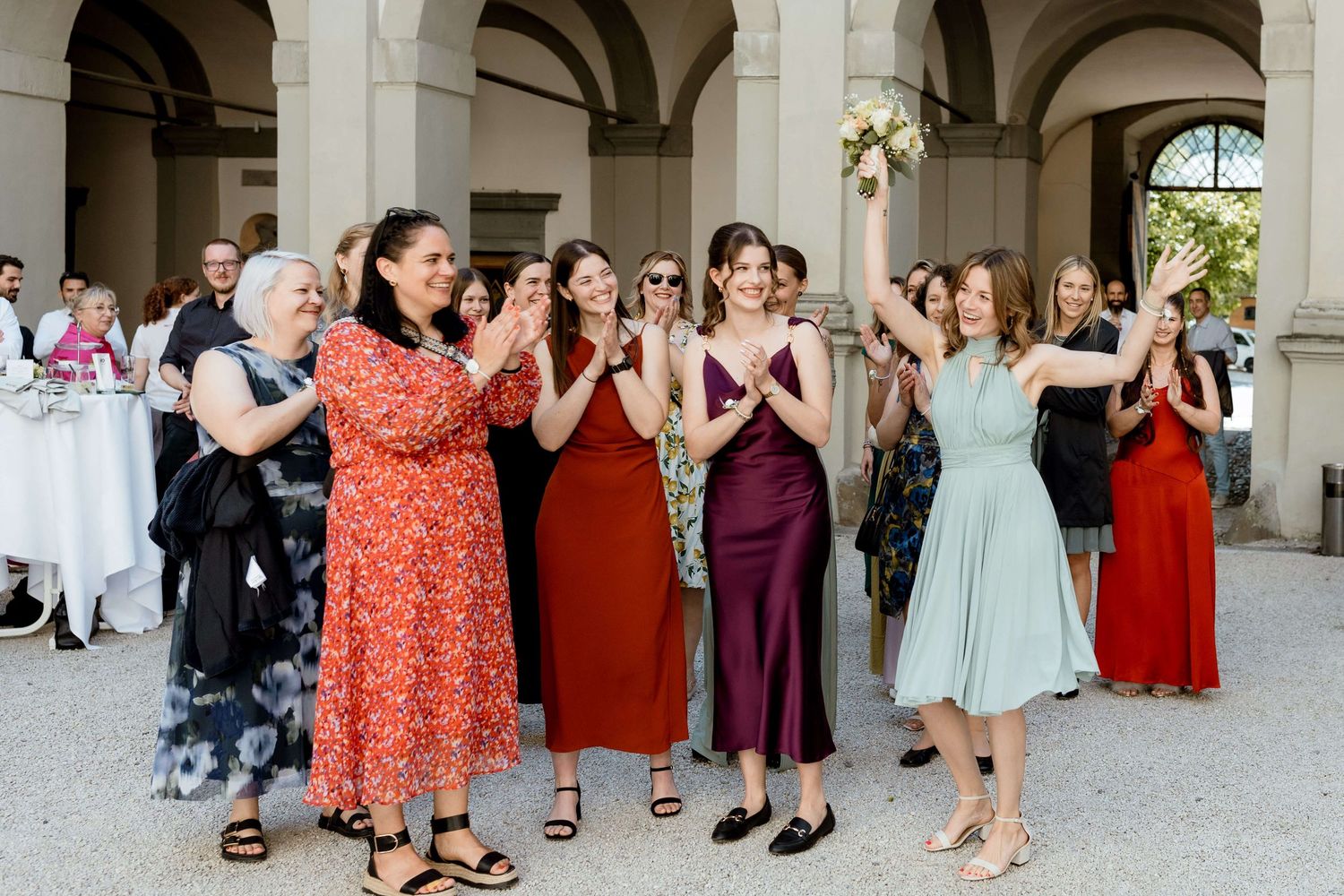 Wedding guests in colorful dresses reaching for bouquet toss at elegant outdoor reception venue.
