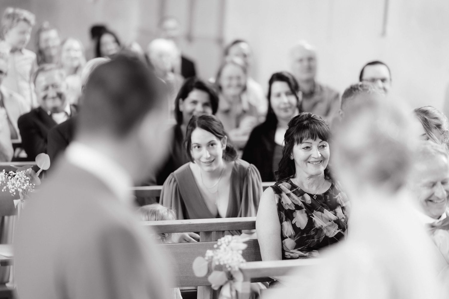Wedding guests smiling and watching ceremony from church pews in black and white photo.
