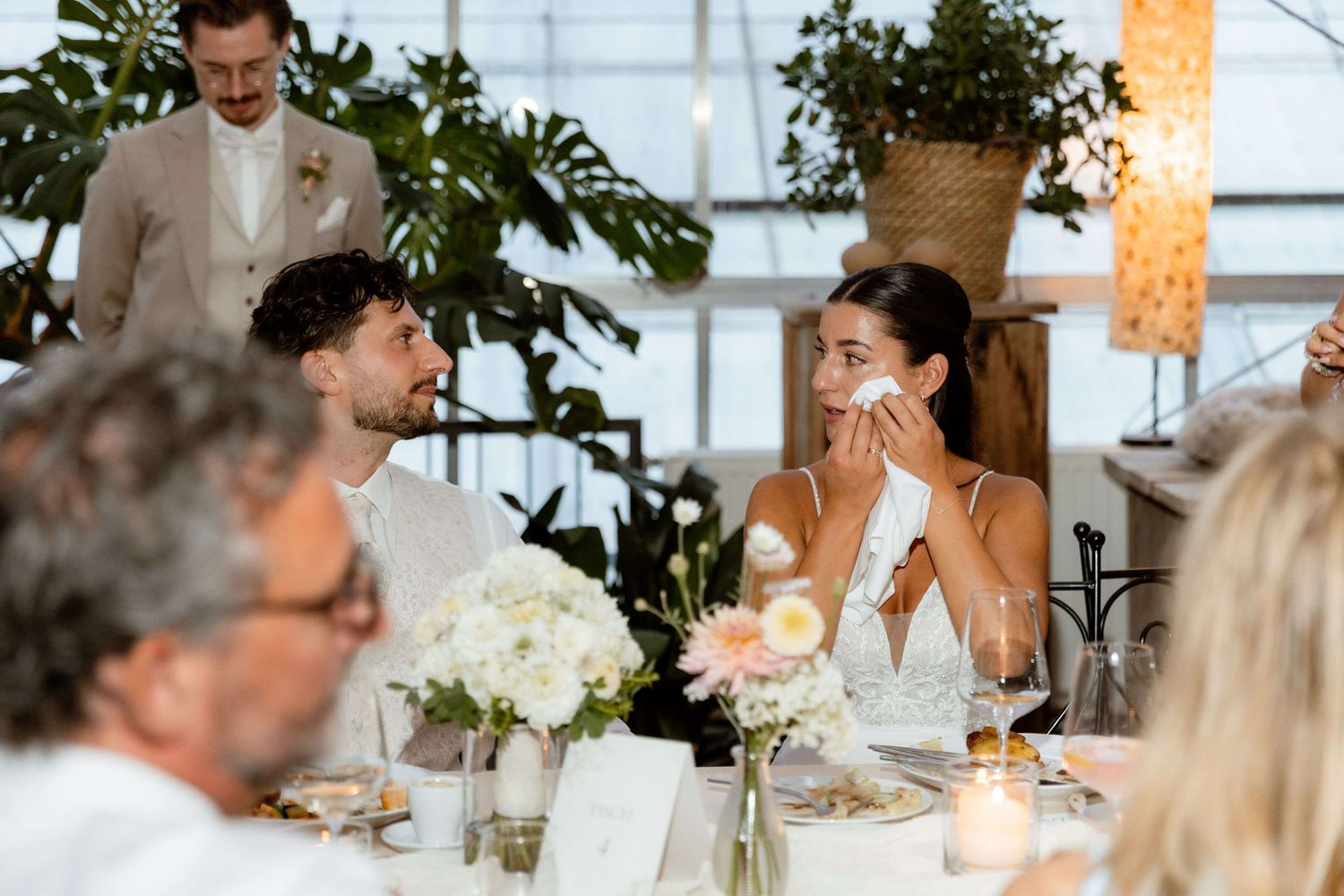 Emotional bride wiping tears at wedding reception table surrounded by floral centerpieces and candles.