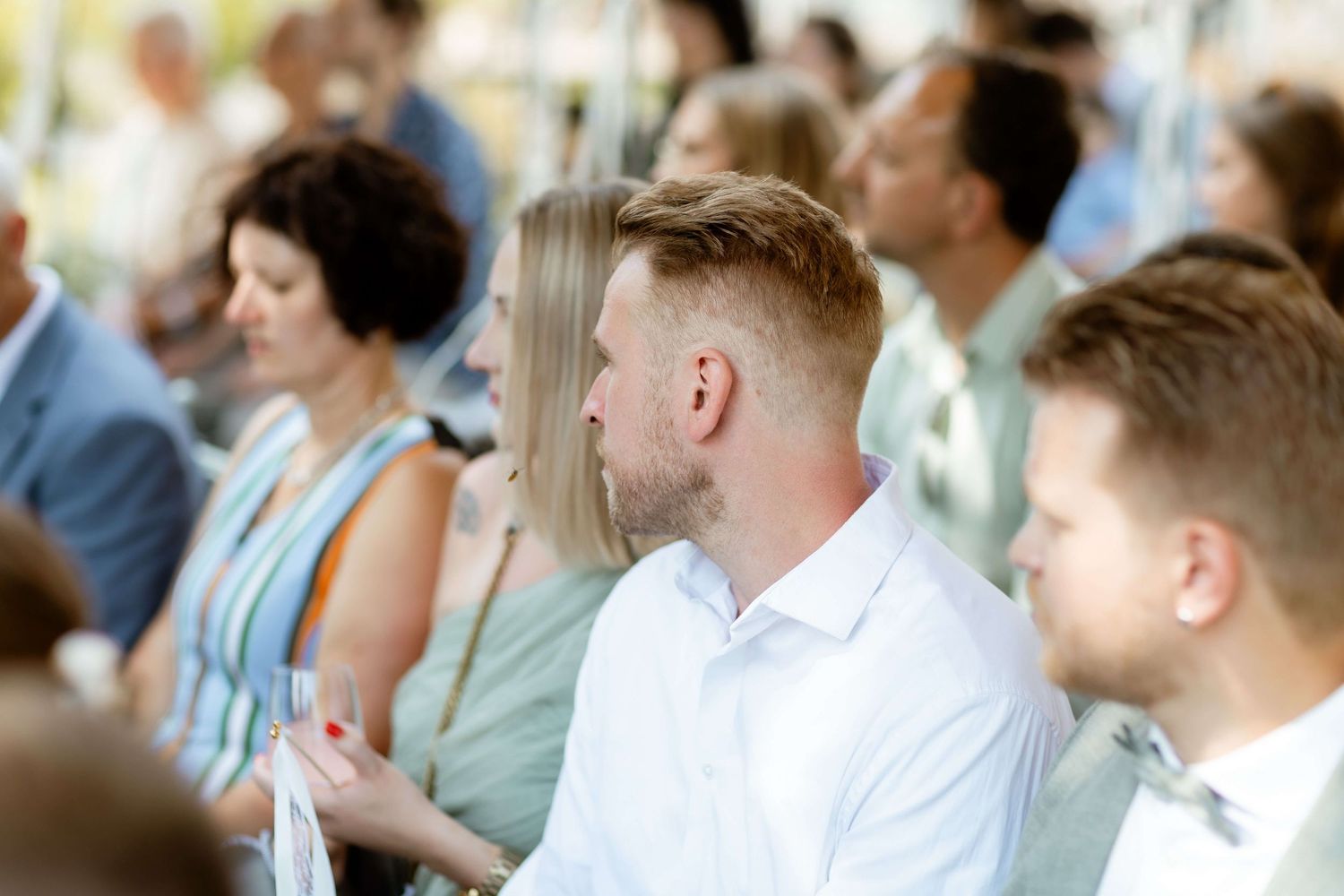 Wedding guests seated outdoors, man in white shirt attentively watching the ceremony.
