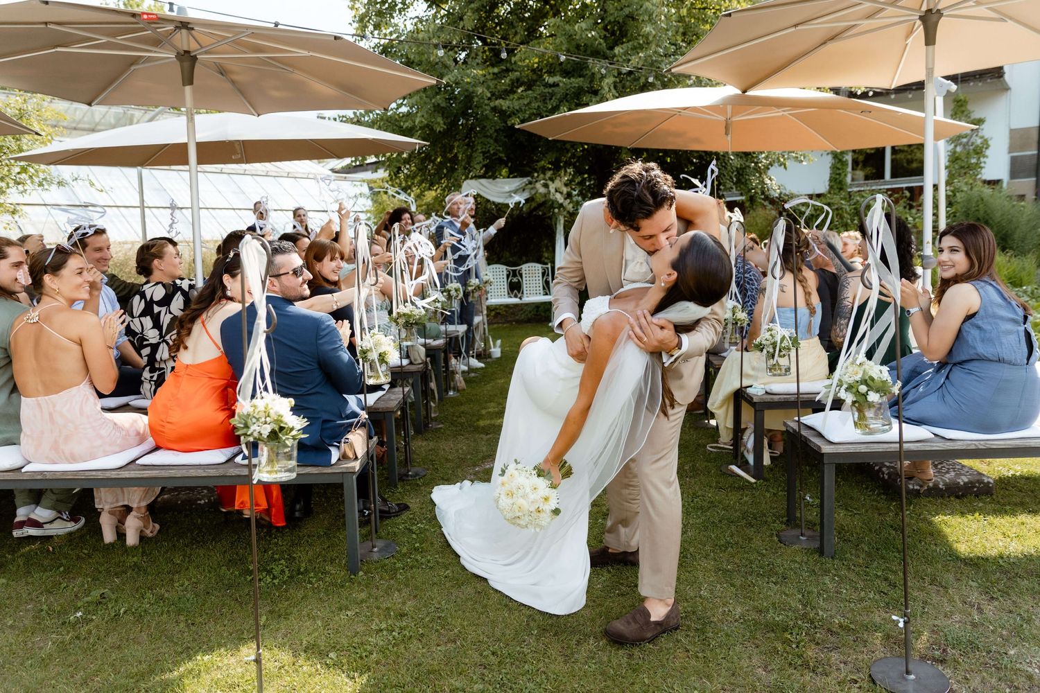 Groom lifting bride during outdoor wedding ceremony with seated guests.