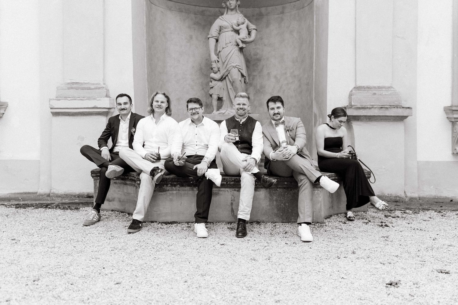 Black and white photo of six wedding guests sitting relaxed on a stone bench near a statue.