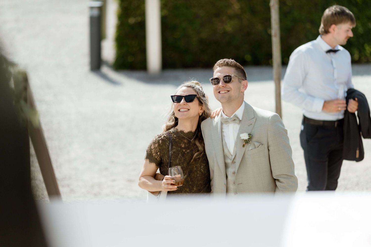Smiling couple in sunglasses poses together outdoors at a sunny wedding reception venue.