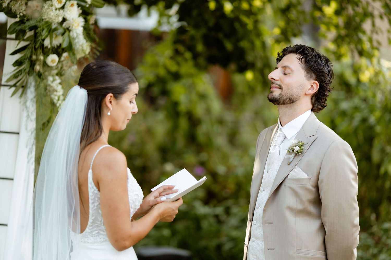 Bride reads vows from booklet to smiling groom in tan suit at outdoor garden wedding.