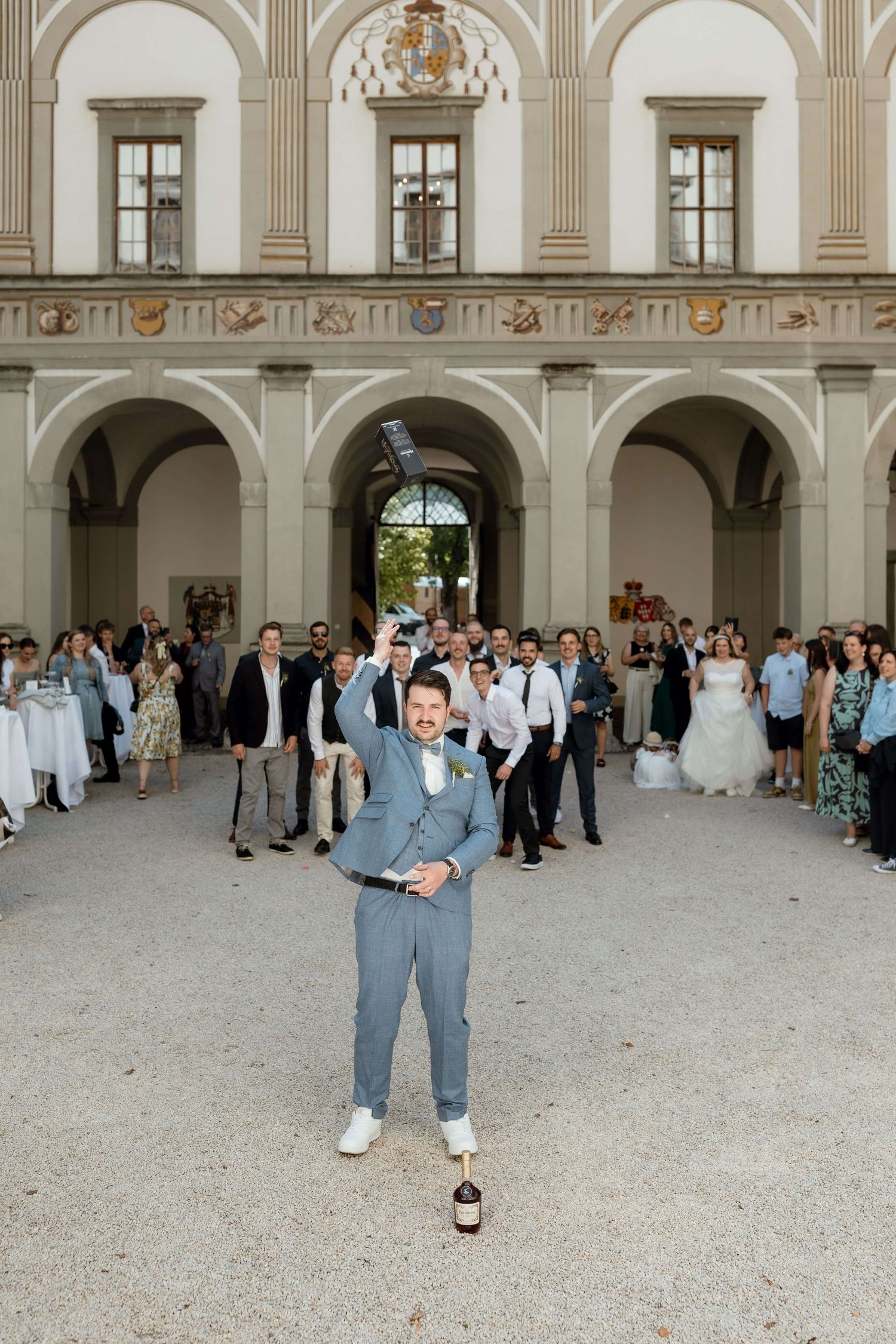 Groom in gray suit tossing garter while guests watch at grand Italian courtyard wedding reception.