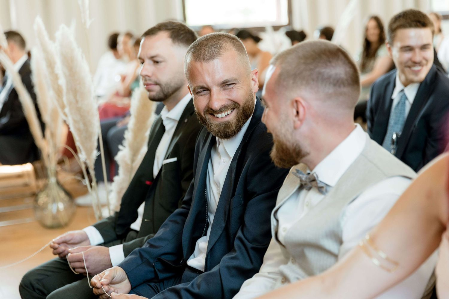 Three groomsmen laughing and chatting joyfully while seated at an indoor wedding ceremony.