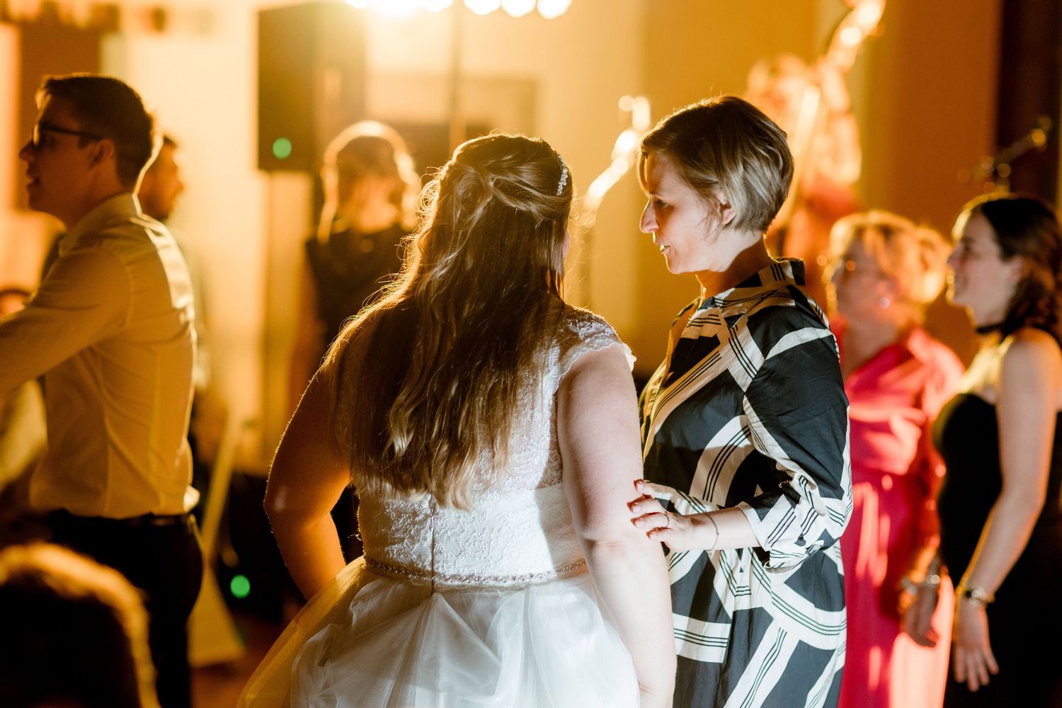 Guests dancing at a wedding reception in warm golden light, with bride in white dress visible.