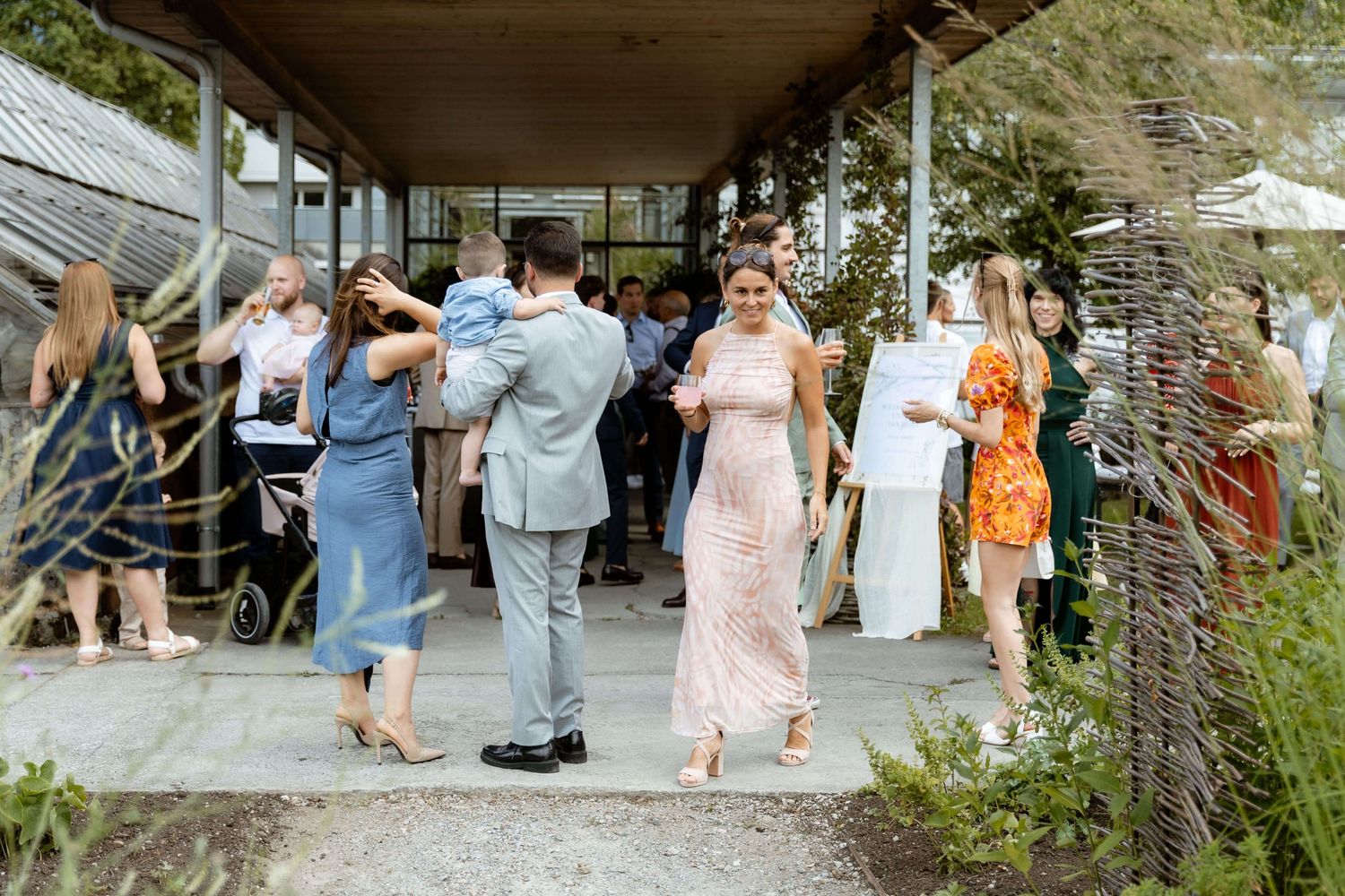 Wedding guests gather and mingle outside a greenhouse venue on a sunny day.