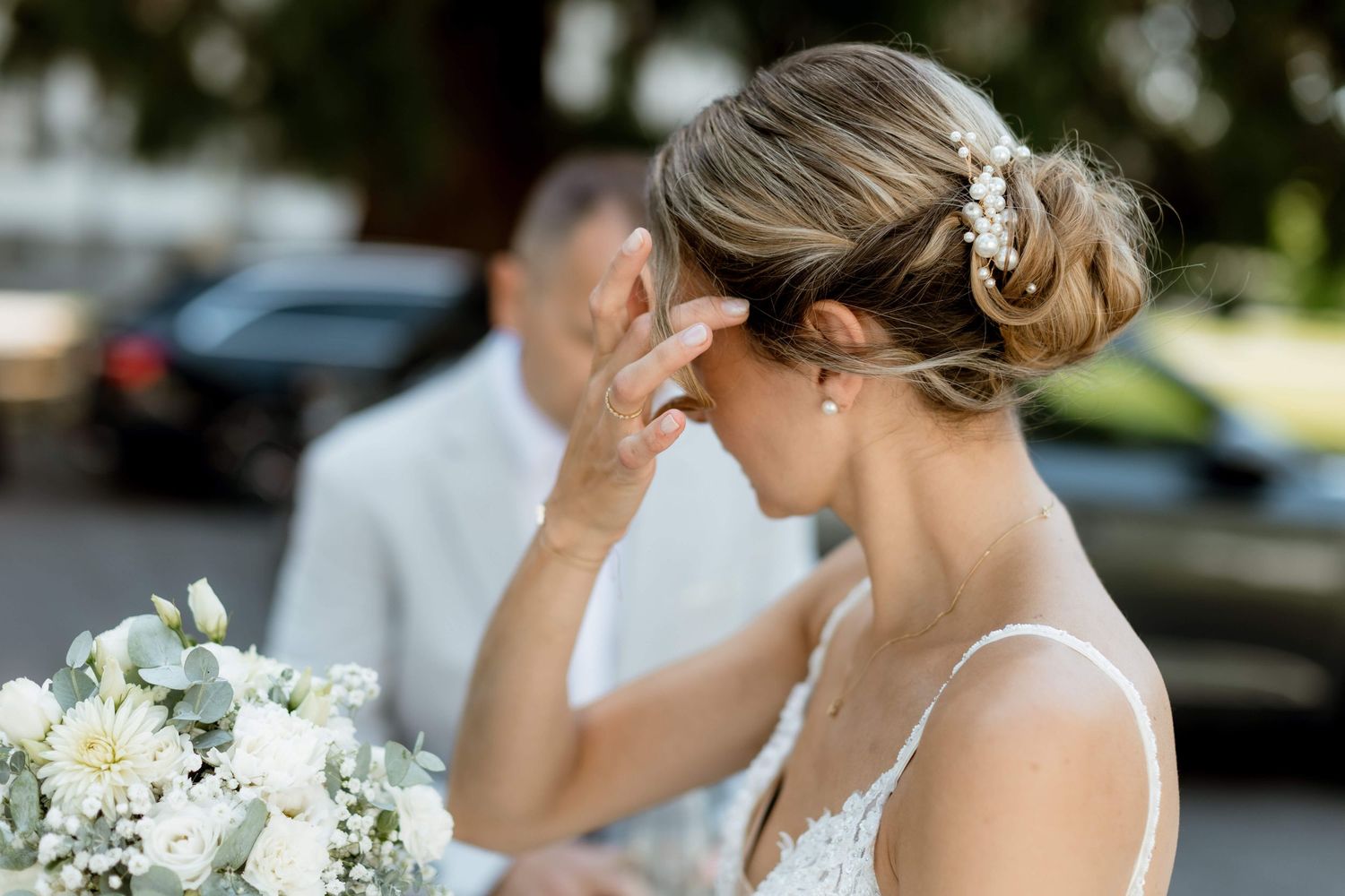 Emotional bride in lace dress touches her face while holding white floral bouquet at wedding ceremony.