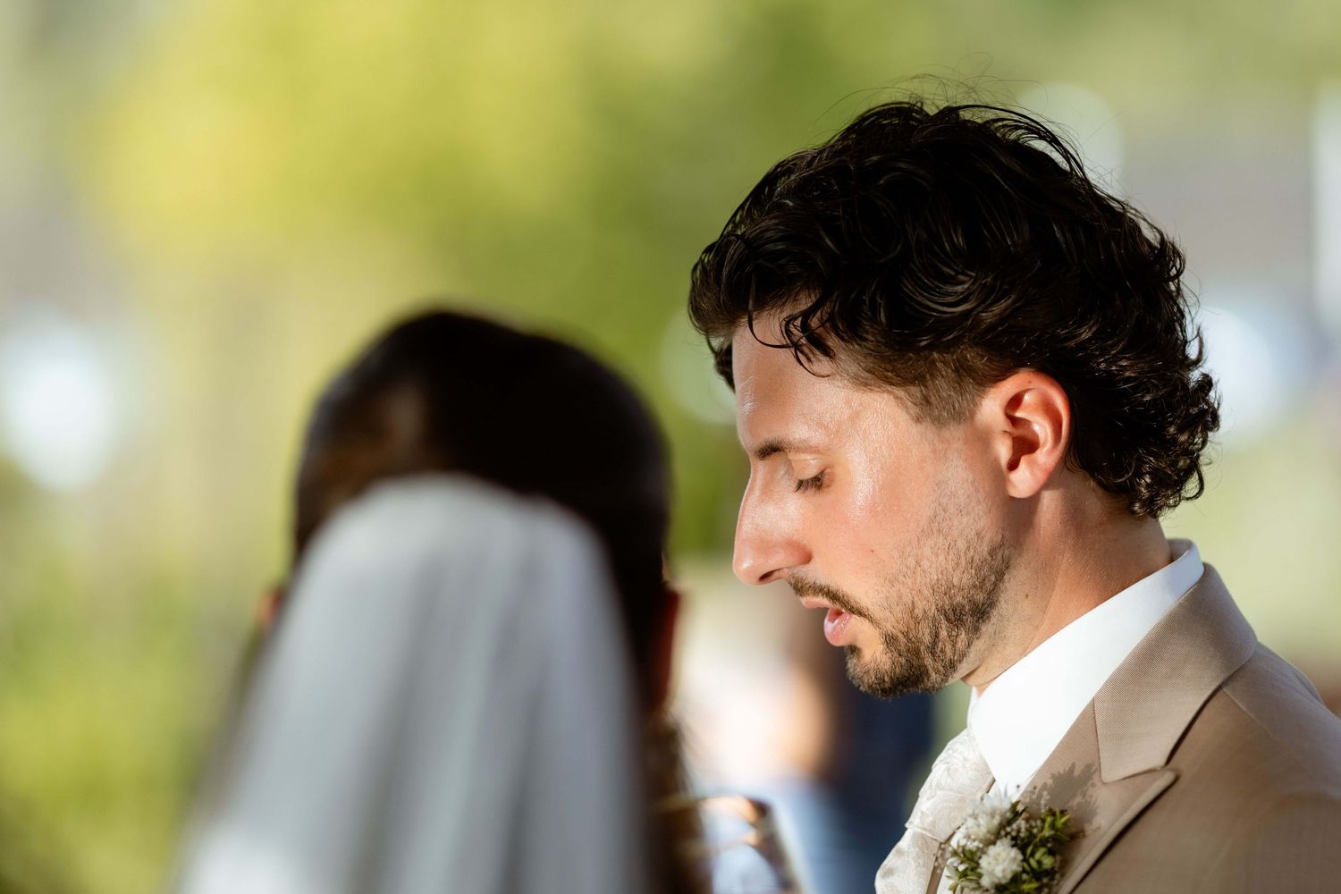 Groom in beige suit with eyes closed during wedding ceremony.