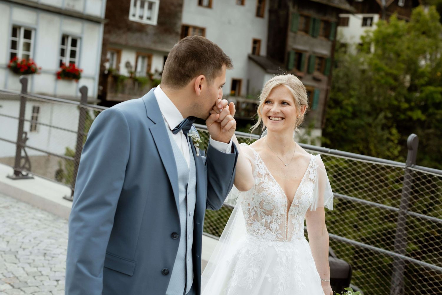 Groom in blue suit kissing bride's hand while she smiles during outdoor wedding photo session.
